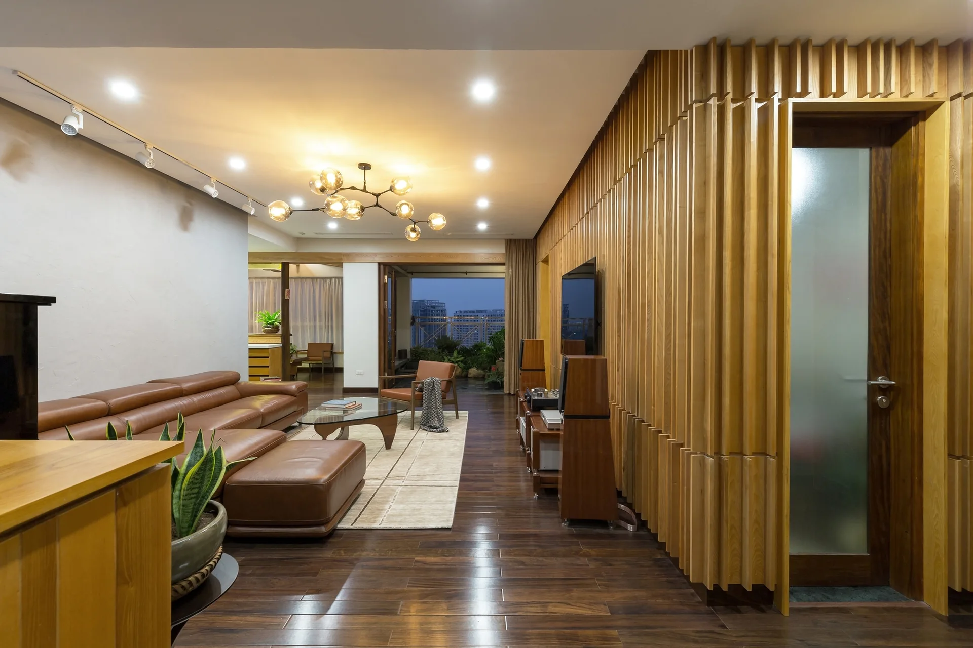 A modern living room features a dark hardwood floor, a large brown leather sofa, and a vertical wood slat wall with a frosted glass door.