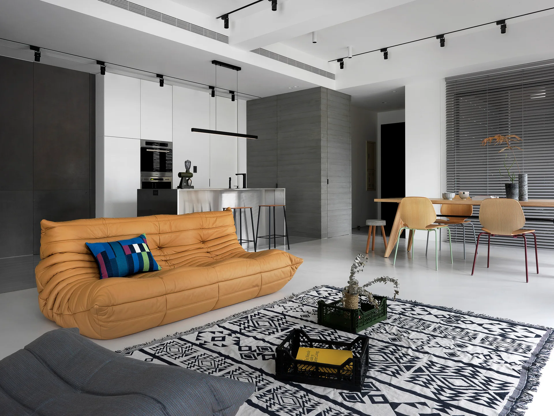 An open-plan room features a tufted mustard yellow sofa on a patterned black and white rug, with a kitchen island and wooden dining table in the background.