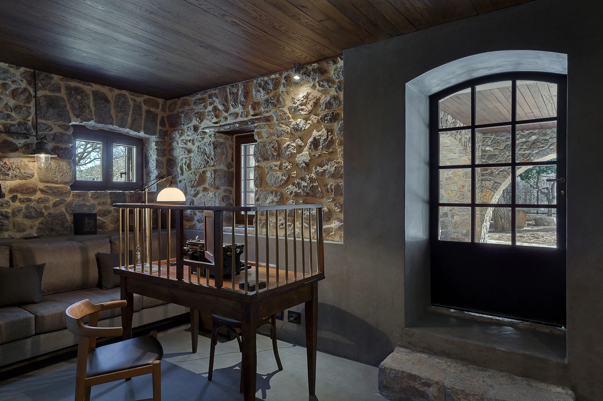 A rustic home office featuring exposed stone walls, a dark wood plank ceiling, a built-in sofa, and a wooden desk with a brass railing.
