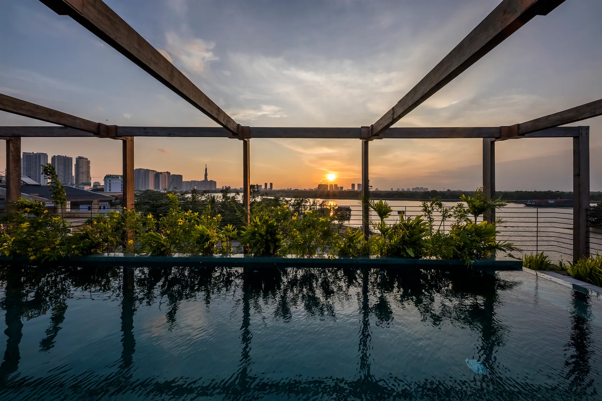 An outdoor rooftop swimming pool with a wooden pergola frame overlooks a river and city skyline during sunset.
