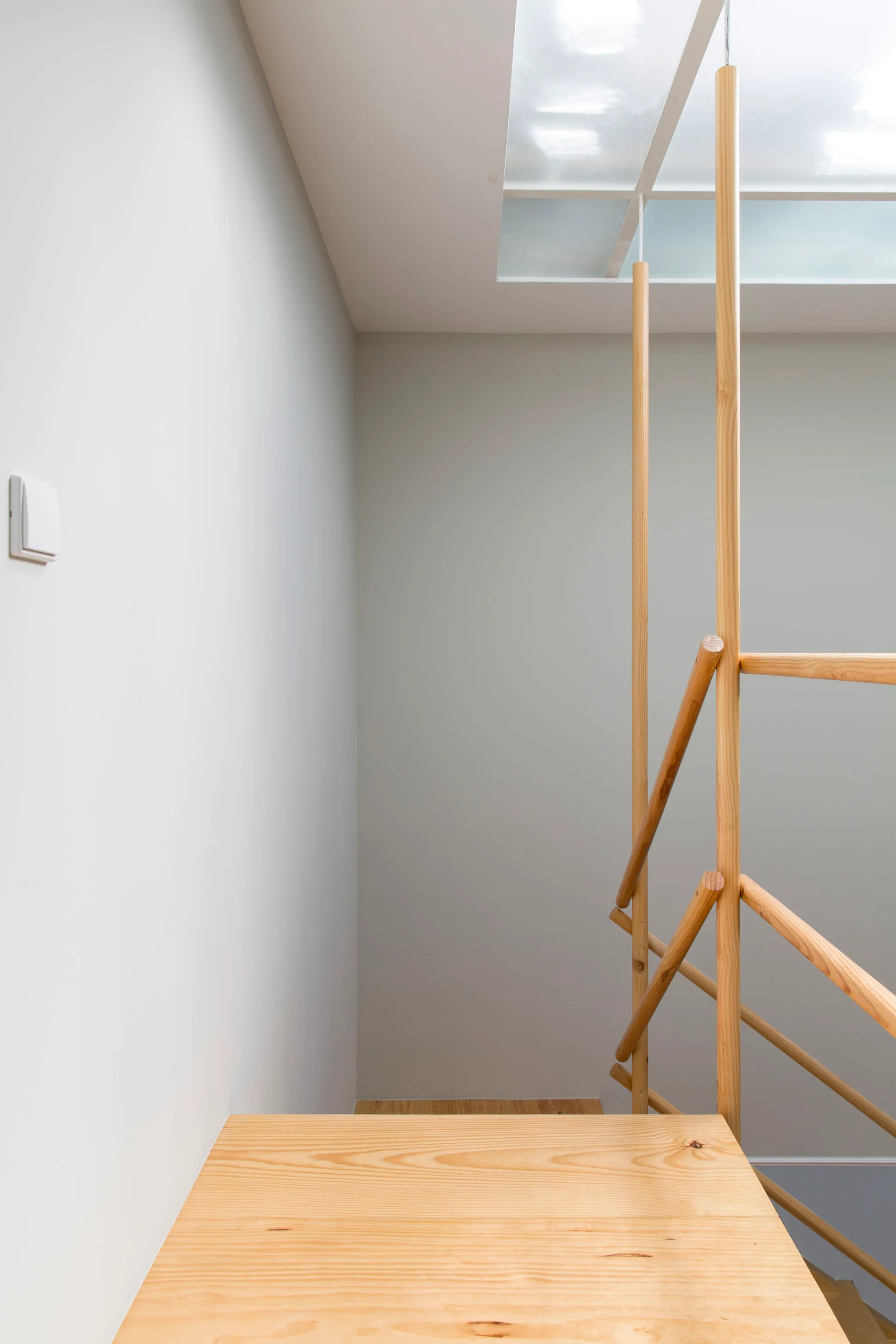 A minimalist wood staircase landing features a light wood railing and steps beneath a bright ceiling skylight.