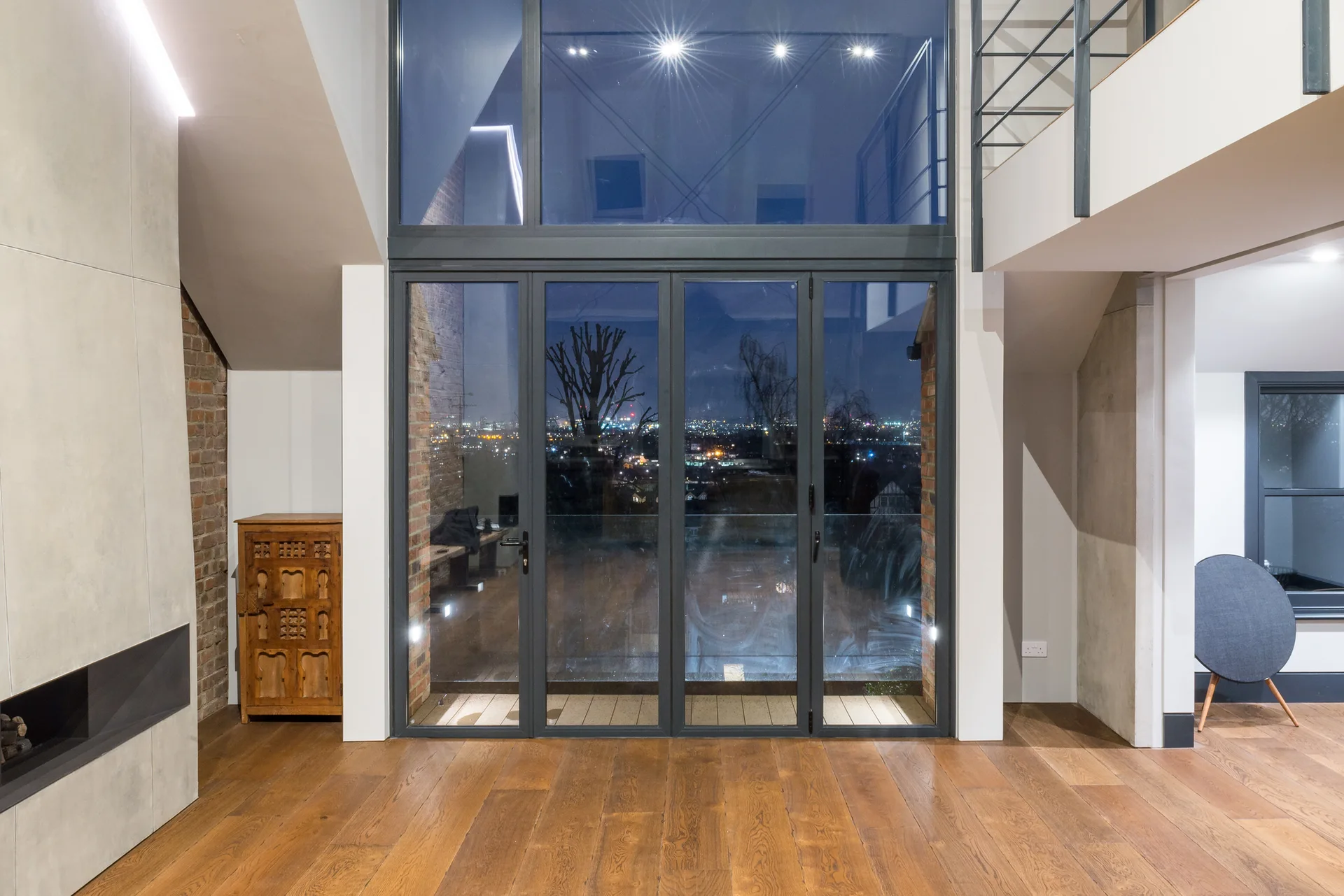 A contemporary double-height room features large dark-framed glass bifold doors, wide-plank hardwood floors, and a small wooden cabinet against a brick wall.