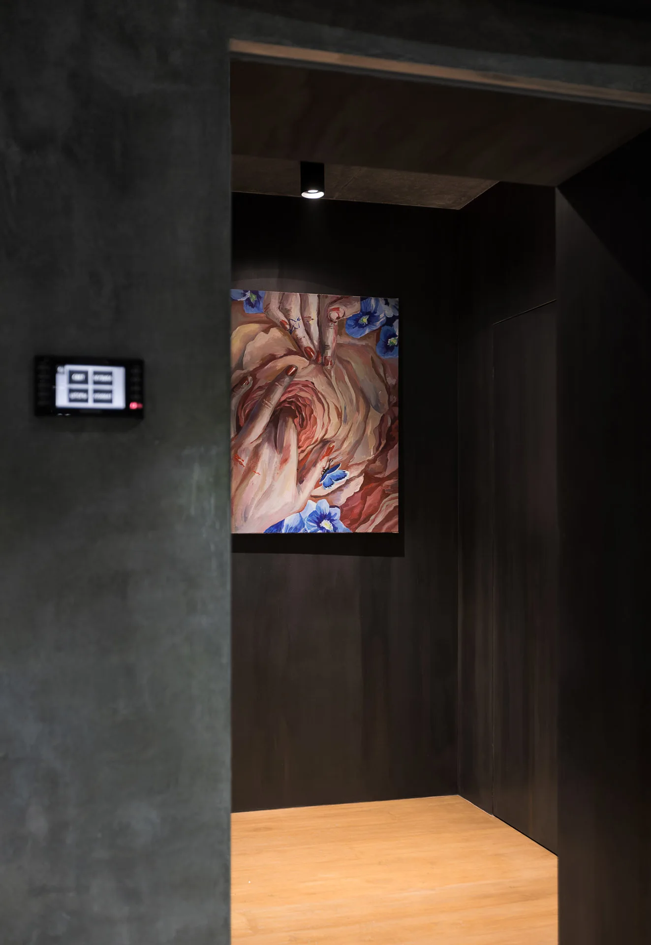 A dark charcoal hallway niche features a figurative painting illuminated by a single ceiling spotlight above warm wood flooring.