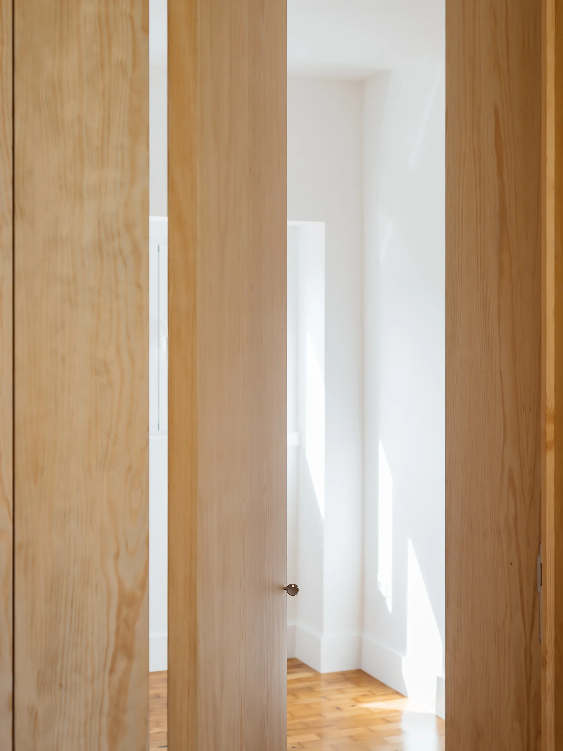 Three vertical natural wood door panels stand partially open, revealing a bright white room with wooden floors and sunlight casting shadows on the wall.