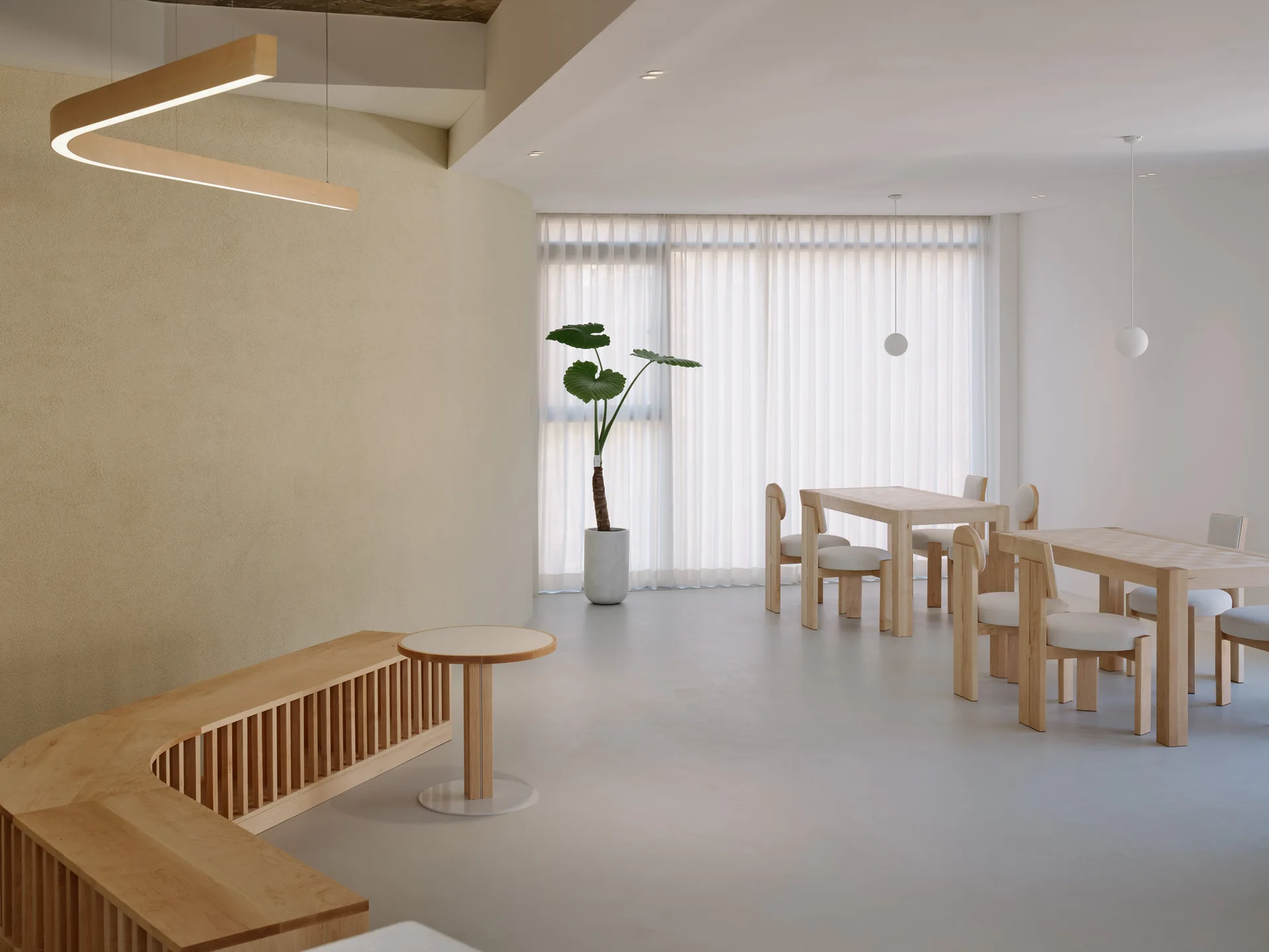 A minimalist dining room features light wood tables and chairs, a curved slatted wooden bench, textured beige walls, and sheer white window curtains.