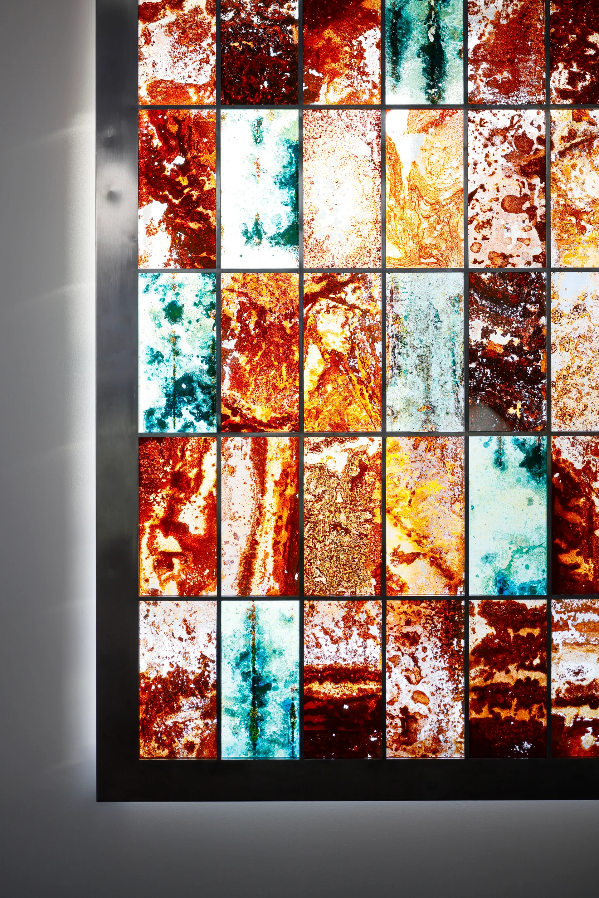 A backlit wall installation featuring a grid of multi-colored textured glass panels in amber, rust, and turquoise framed by brushed metal.