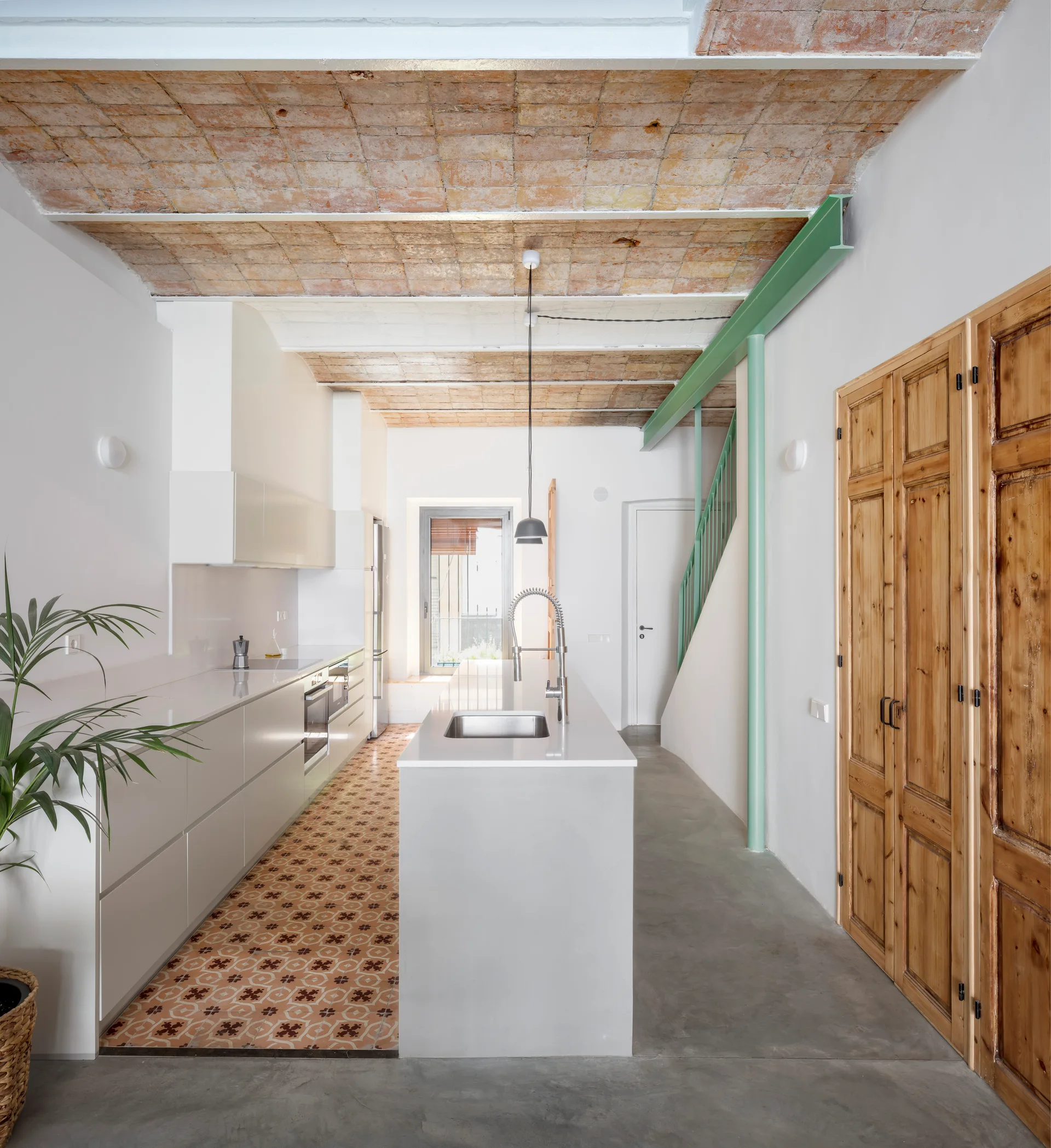 A minimalist modern kitchen with a concrete floor, inlaid patterned tiles, white cabinets, and an exposed brick vaulted ceiling.