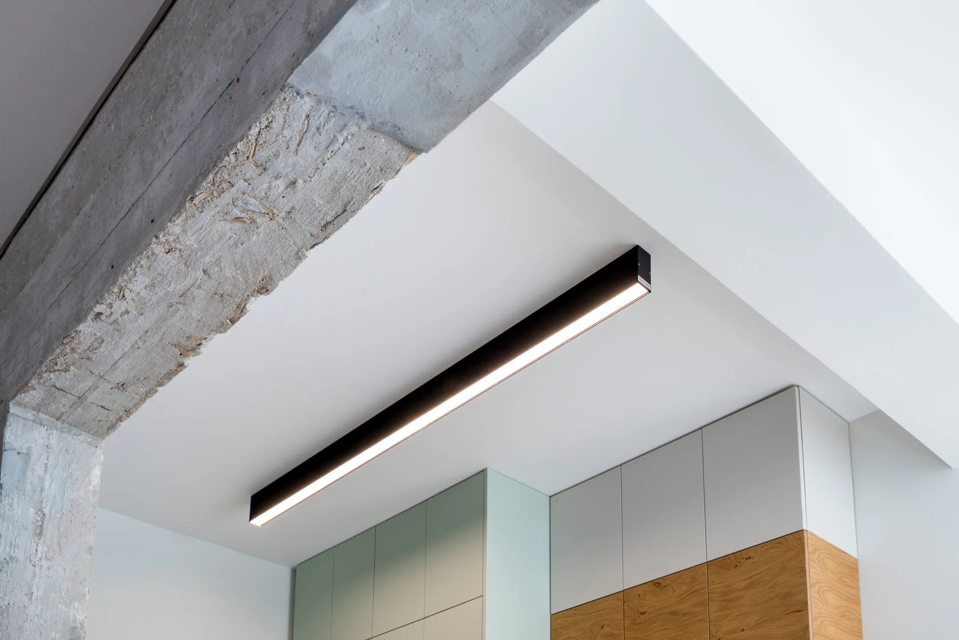 A black linear light fixture is mounted on a white ceiling next to an exposed concrete beam and modern kitchen cabinets.