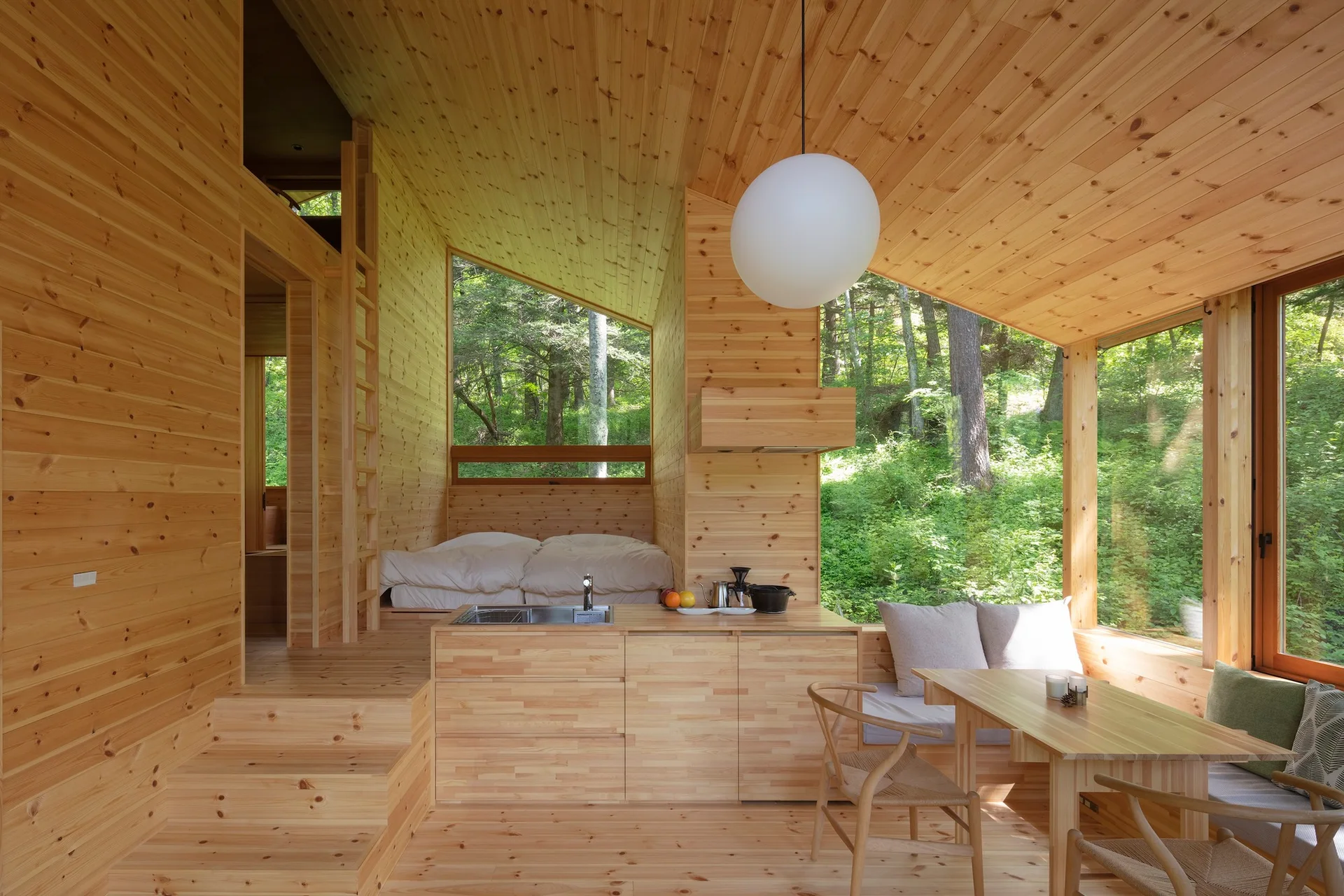 A minimalist cabin interior made entirely of light wood, featuring a kitchenette, built-in window seating, and a raised sleeping area.