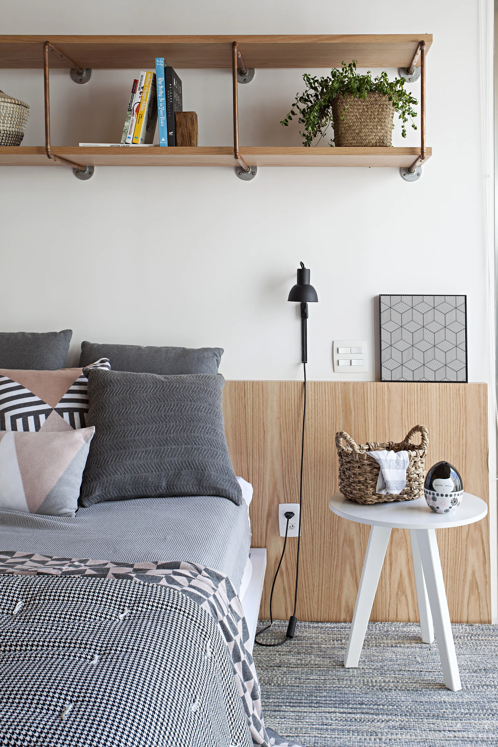 A light wood paneled headboard wall in a bedroom features a black wall sconce, white side table, grey bedding, and industrial pipe shelving above.