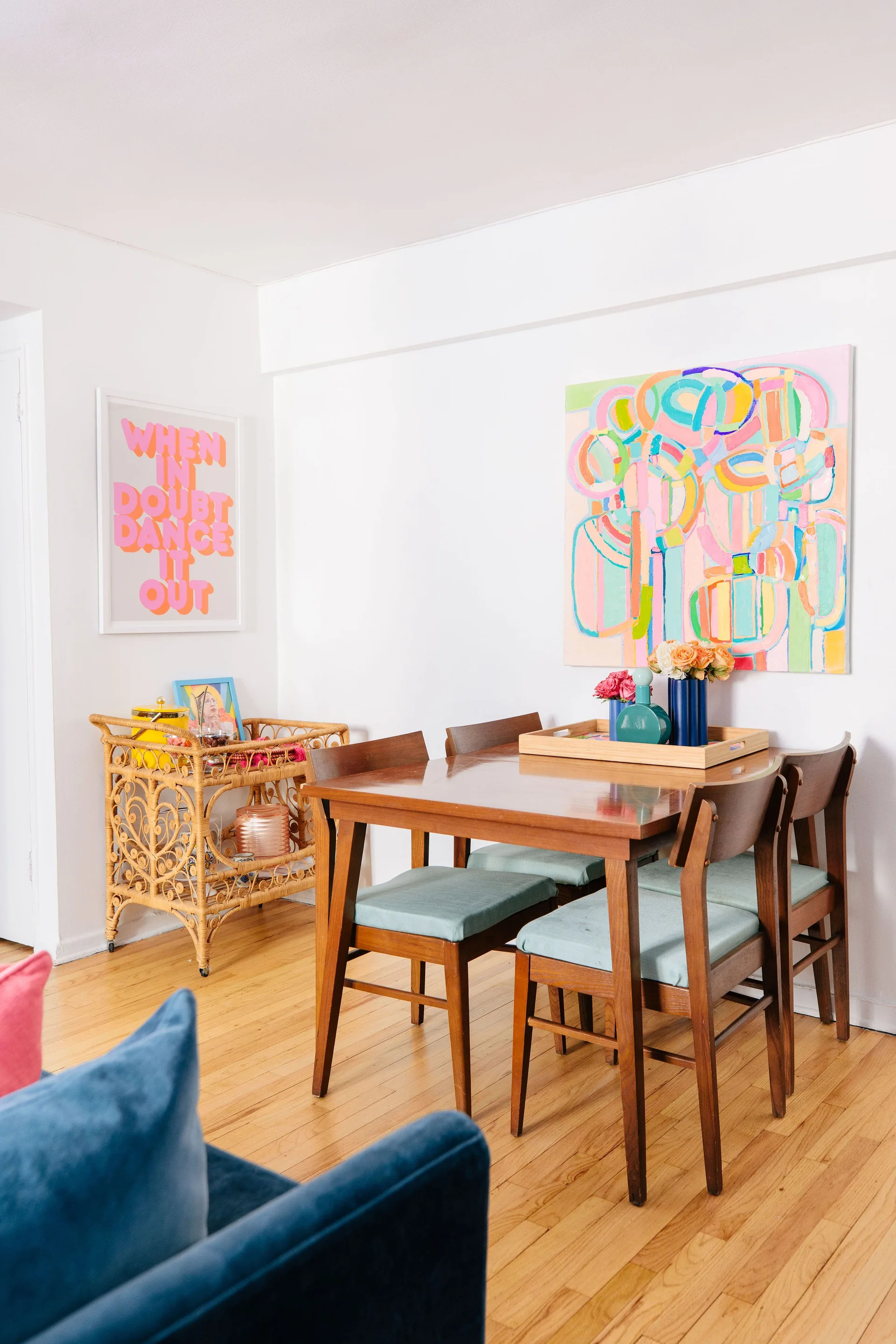 A wooden dining table and chairs with teal cushions sit on a hardwood floor beneath a colorful abstract painting next to a vintage rattan bar cart.