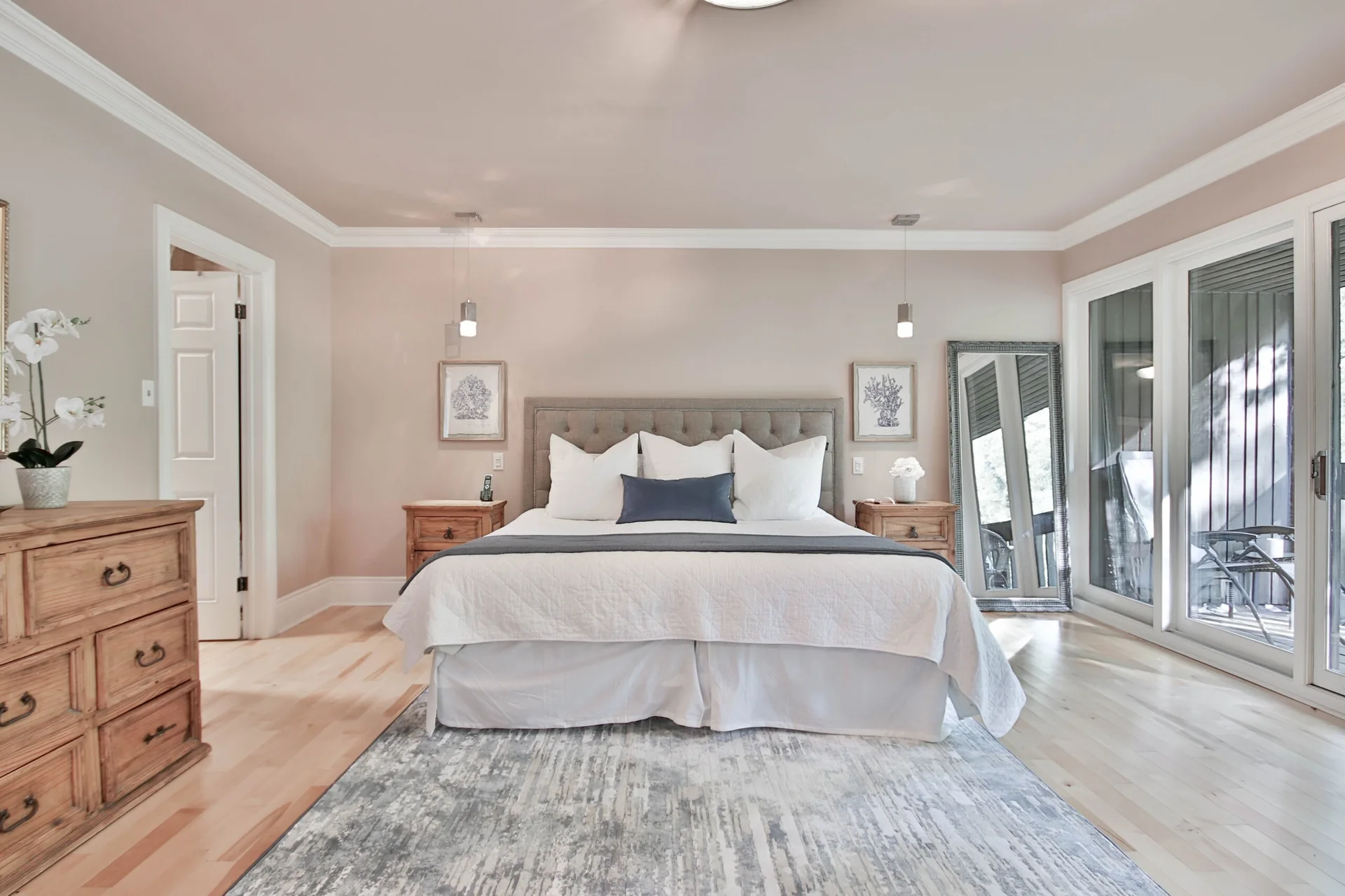 A modern transitional bedroom with soft blush pink walls, an upholstered grey headboard, light wood dresser, and a distressed blue-grey area rug.