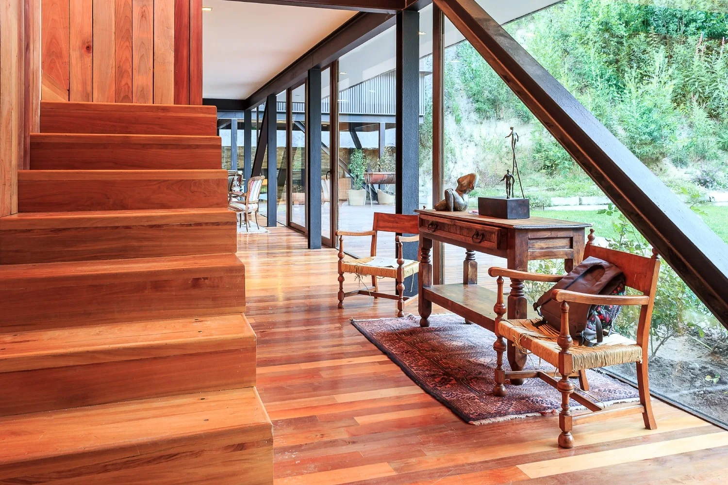 A bright interior hallway features a prominent wooden staircase next to a glass wall showing an outdoor garden.