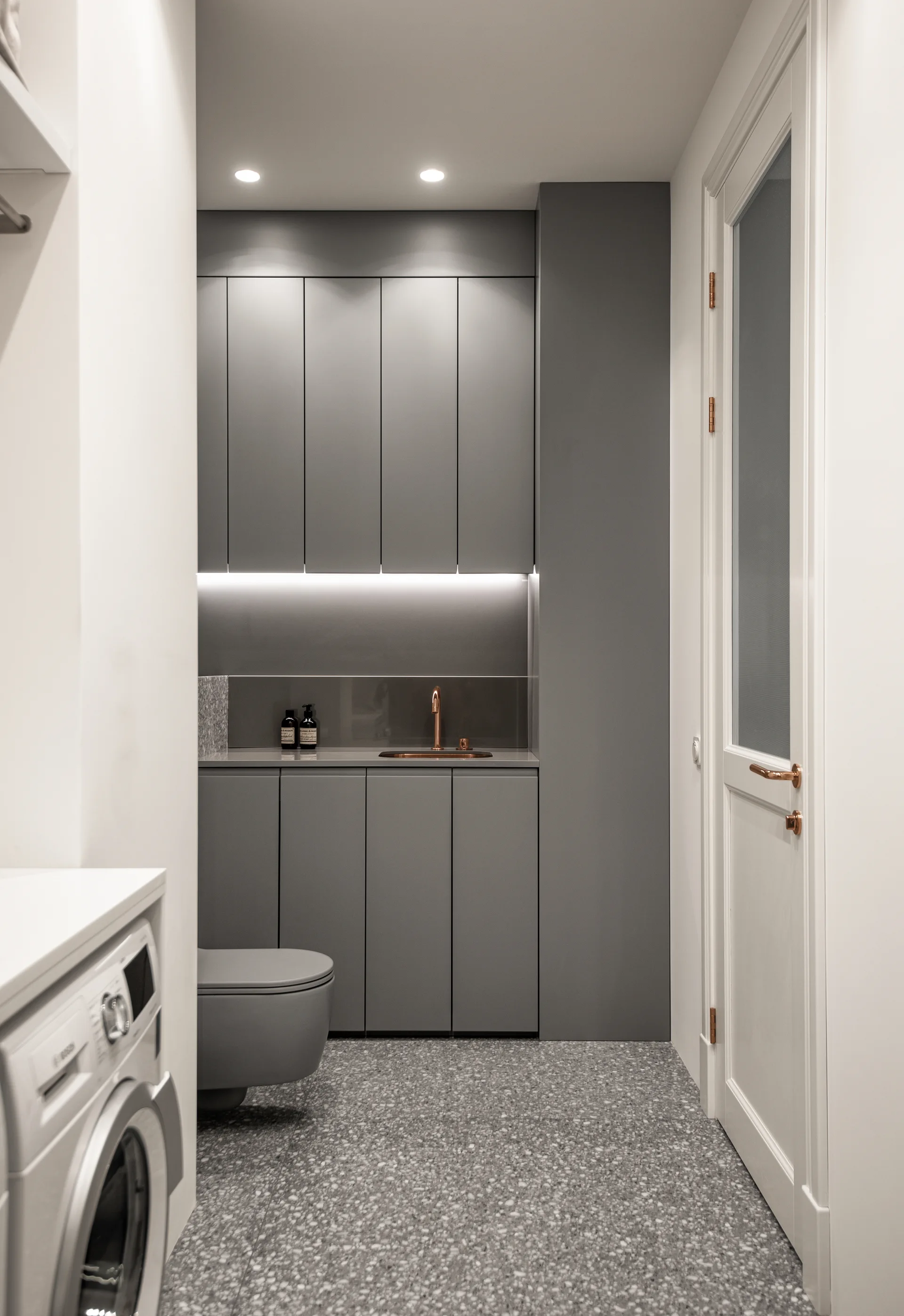 A minimalist laundry room featuring gray flat-panel cabinets, a copper faucet, a terrazzo floor, and a white washing machine.