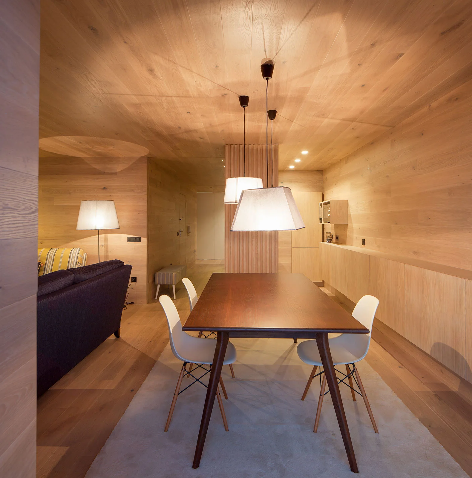 A modern dining area features a dark wood table with white chairs, set against light wood paneled walls and ceiling with glowing pendant lights.