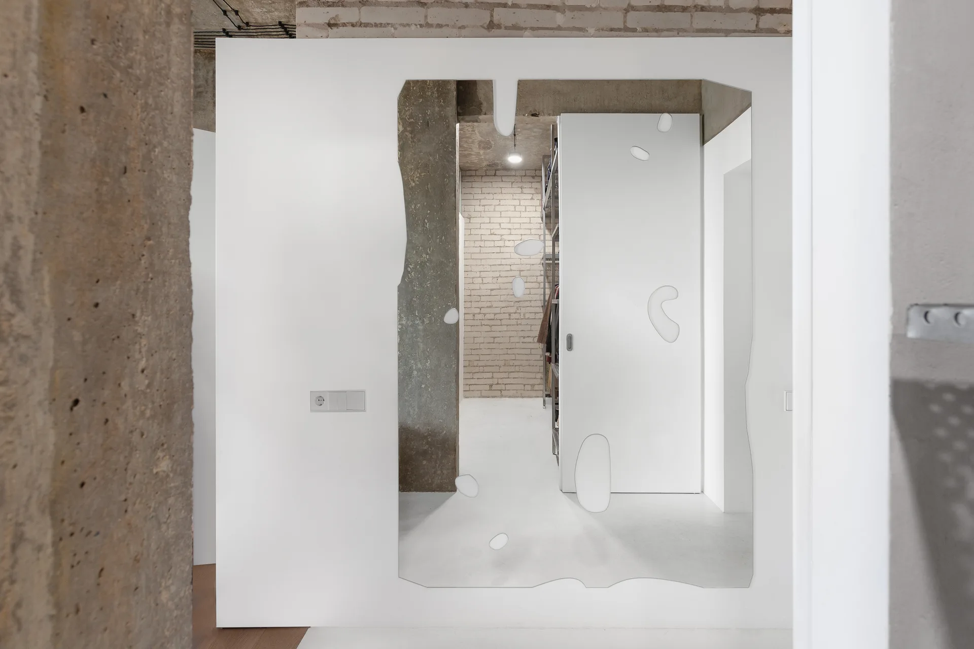 An irregularly shaped mirror with droplet cutouts mounted on a white wall reflects a walk in closet with brick and raw concrete.