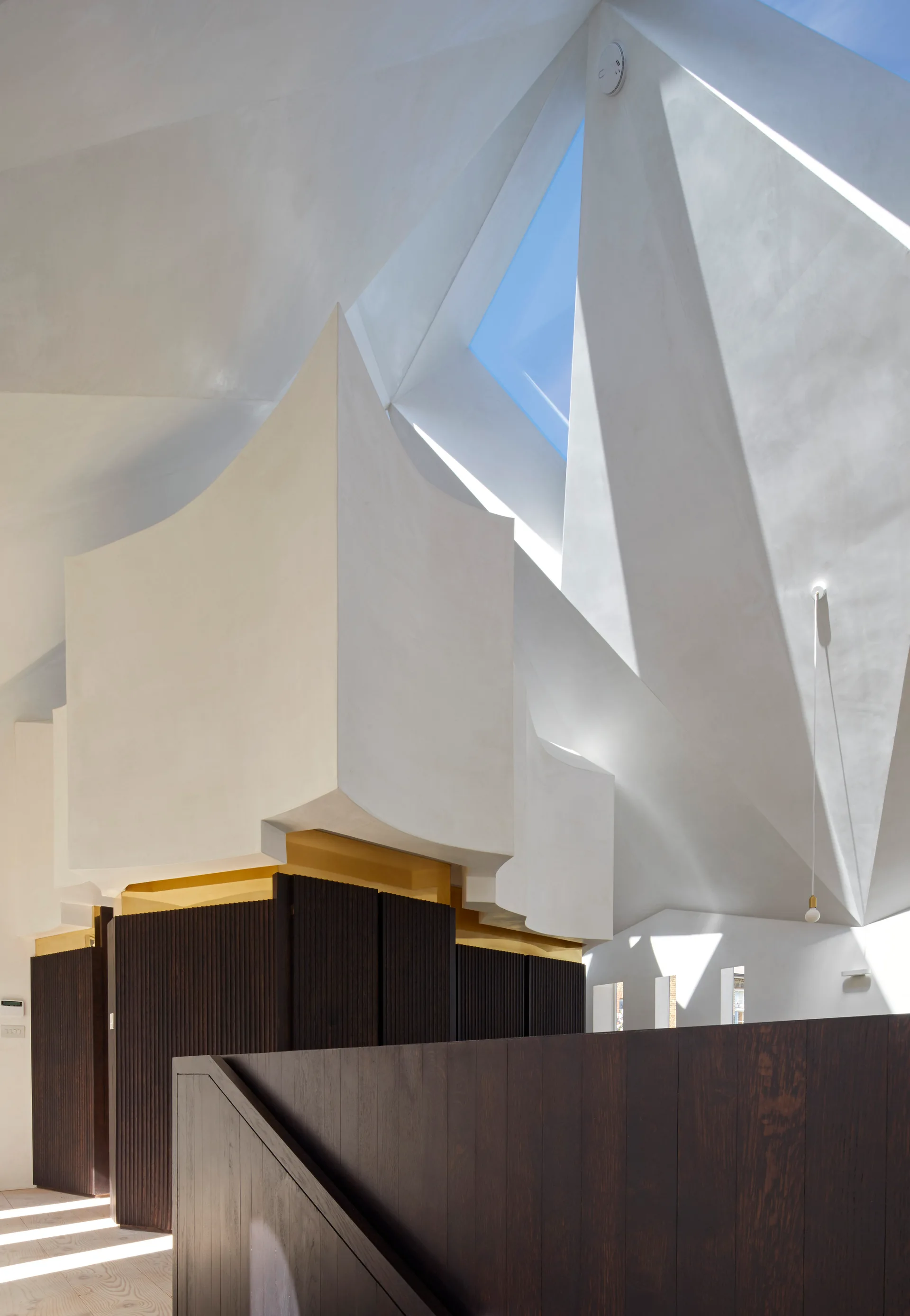 A sculptural white plaster ceiling with a triangular skylight sits above dark wood slatted wall paneling and a dark wood banister.