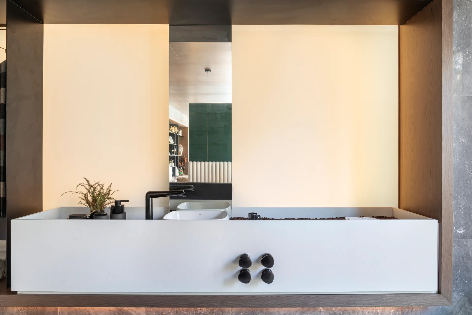 A minimalist bathroom vanity featuring a wide white stone sink basin, a vertical mirror, warm backlighting, and matte black fixtures.