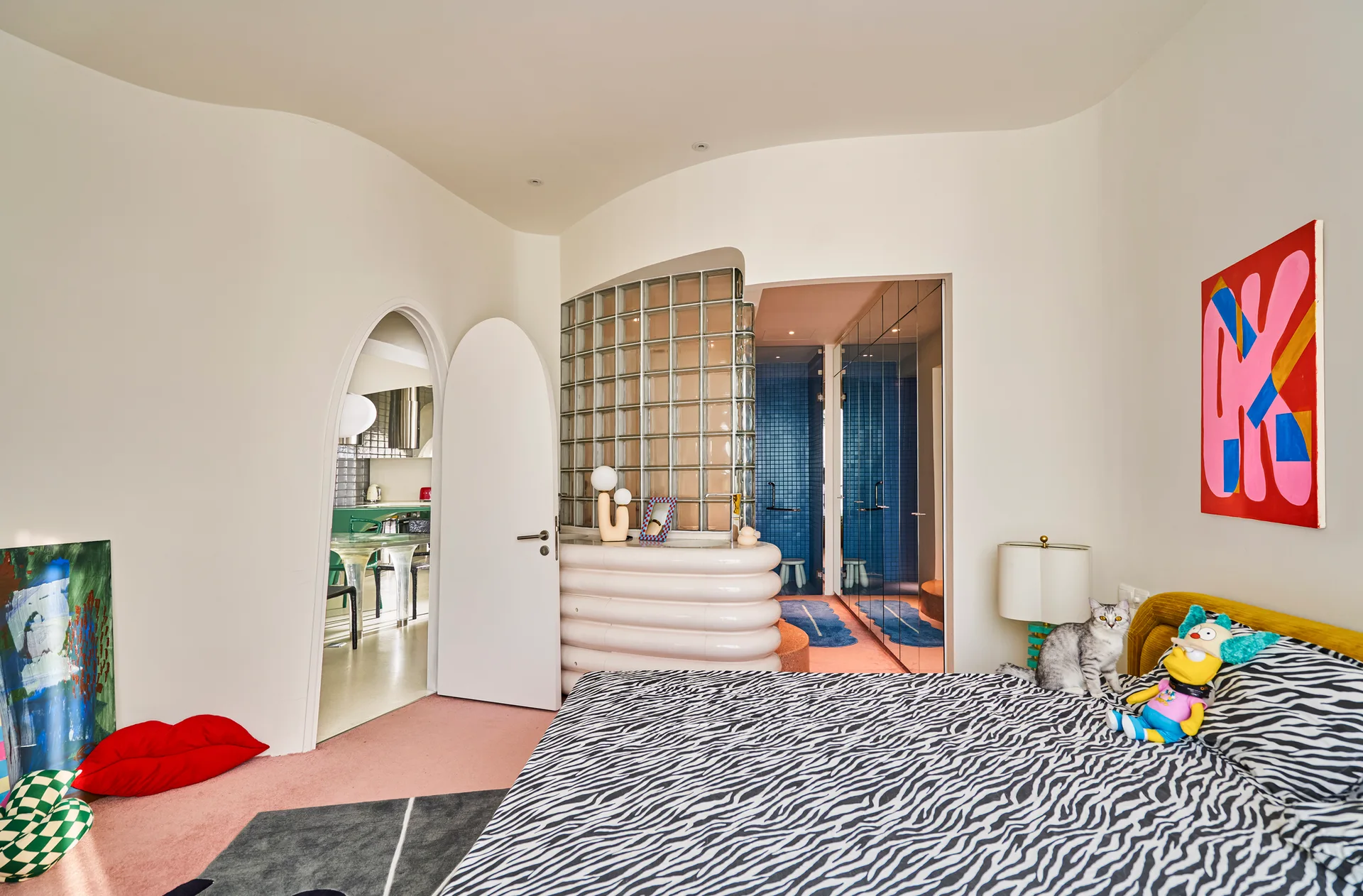 An eclectic bedroom features an arched white doorway, a glass block partition wall, a curved pink console, and a bed with zebra-print sheets.
