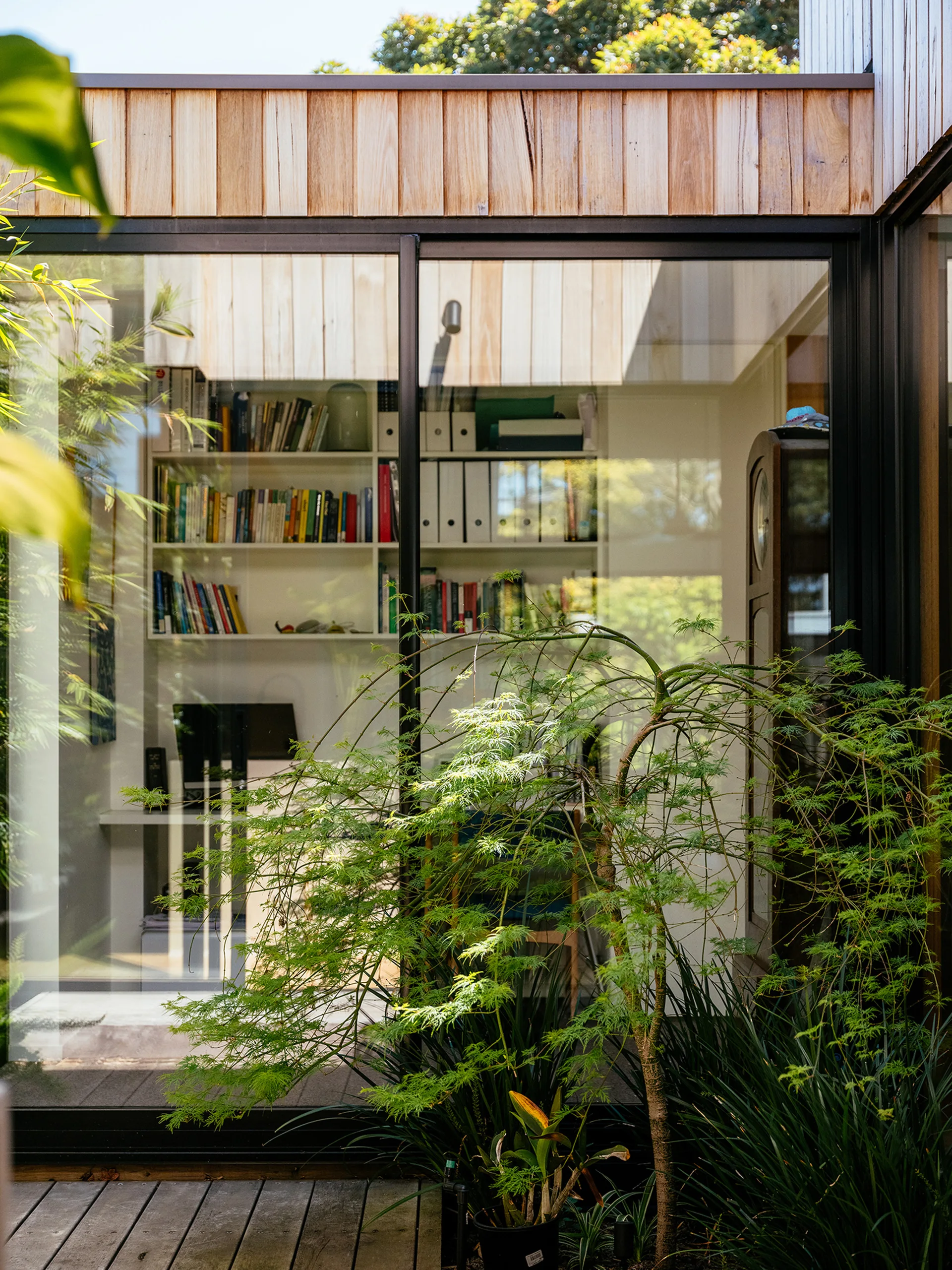 A view through a large glass sliding door from a wooden deck showing a home office filled with white bookshelves and a desk.
