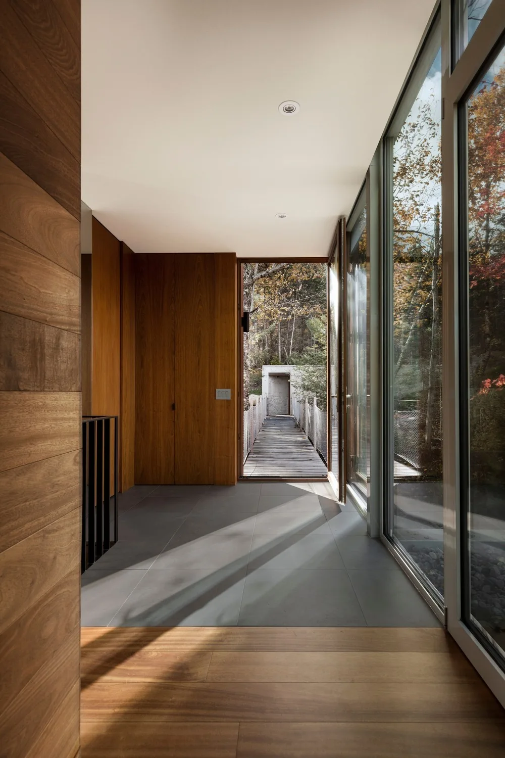 A modern hallway features warm wood-paneled walls, gray stone floor tiles, and large glass doors looking out to an exterior wooden walkway.