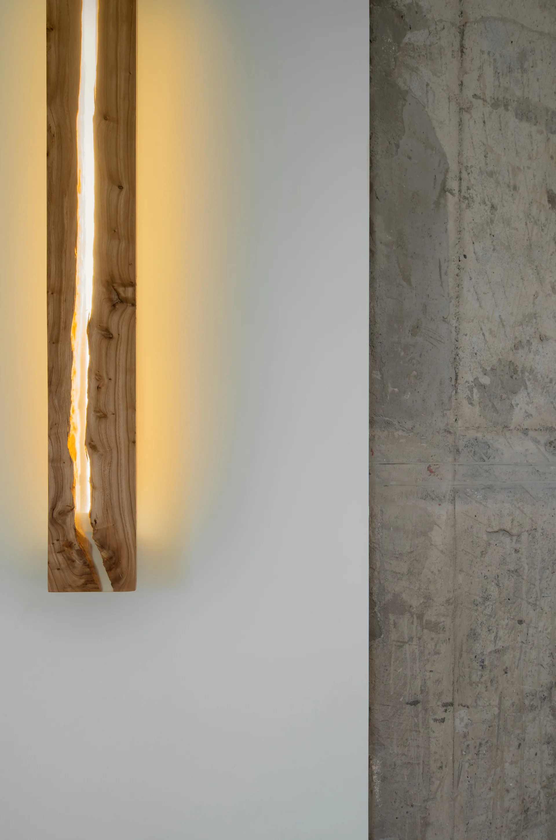 A vertical wooden wall sconce with a warm glowing light strip sits on a smooth white wall next to a raw concrete surface.