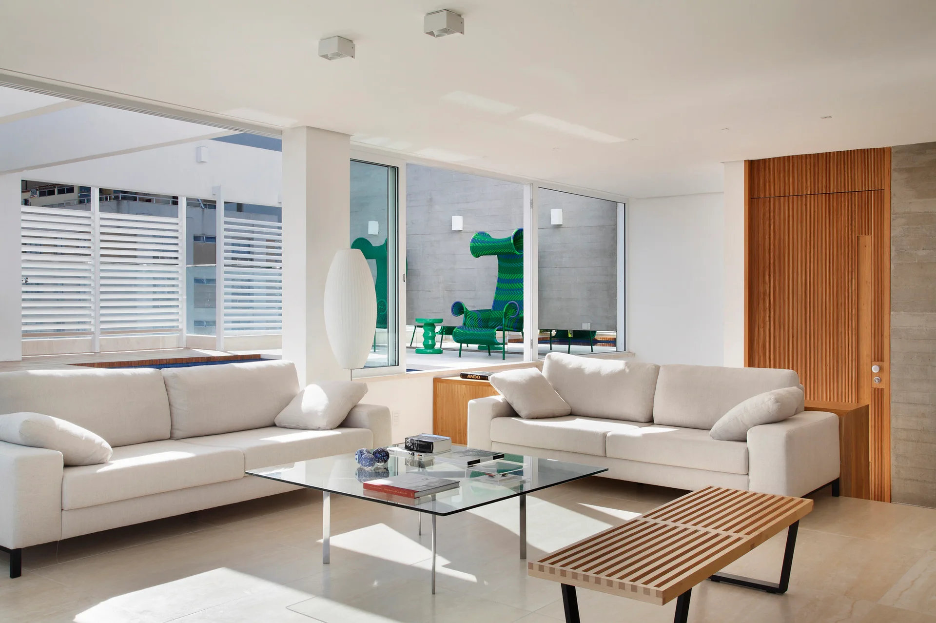 A bright living room features two cream sofas, a glass coffee table, a slatted wood bench, and glass doors leading to an outdoor patio.