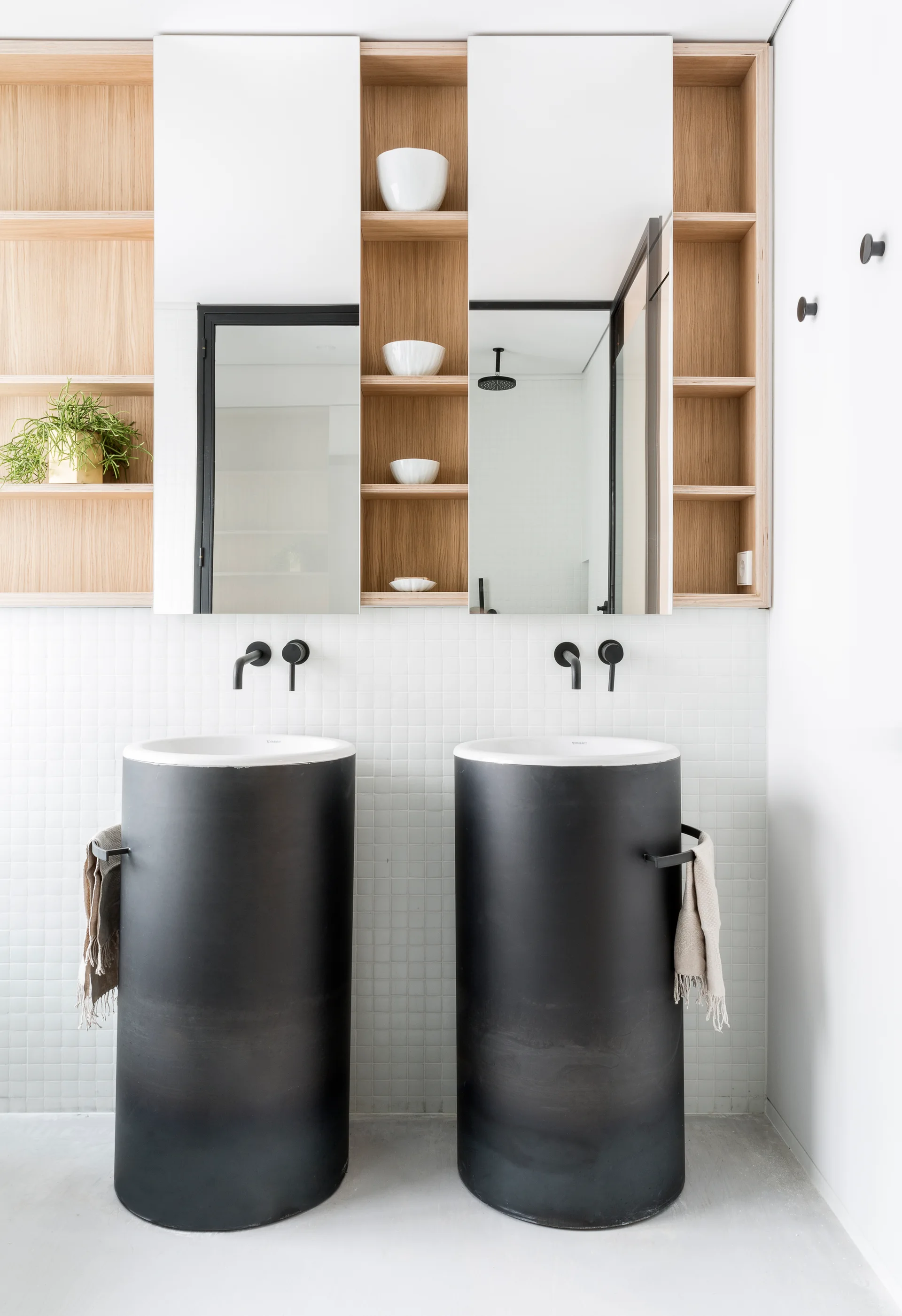 A modern bathroom features two black cylindrical pedestal sinks set against a white square mosaic tile wall with integrated light wood shelves.