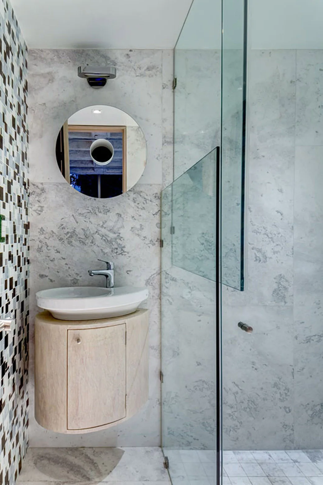 A modern bathroom features a wall-mounted curved wood vanity with a white vessel sink, a round mirror, and large marble wall tiles.