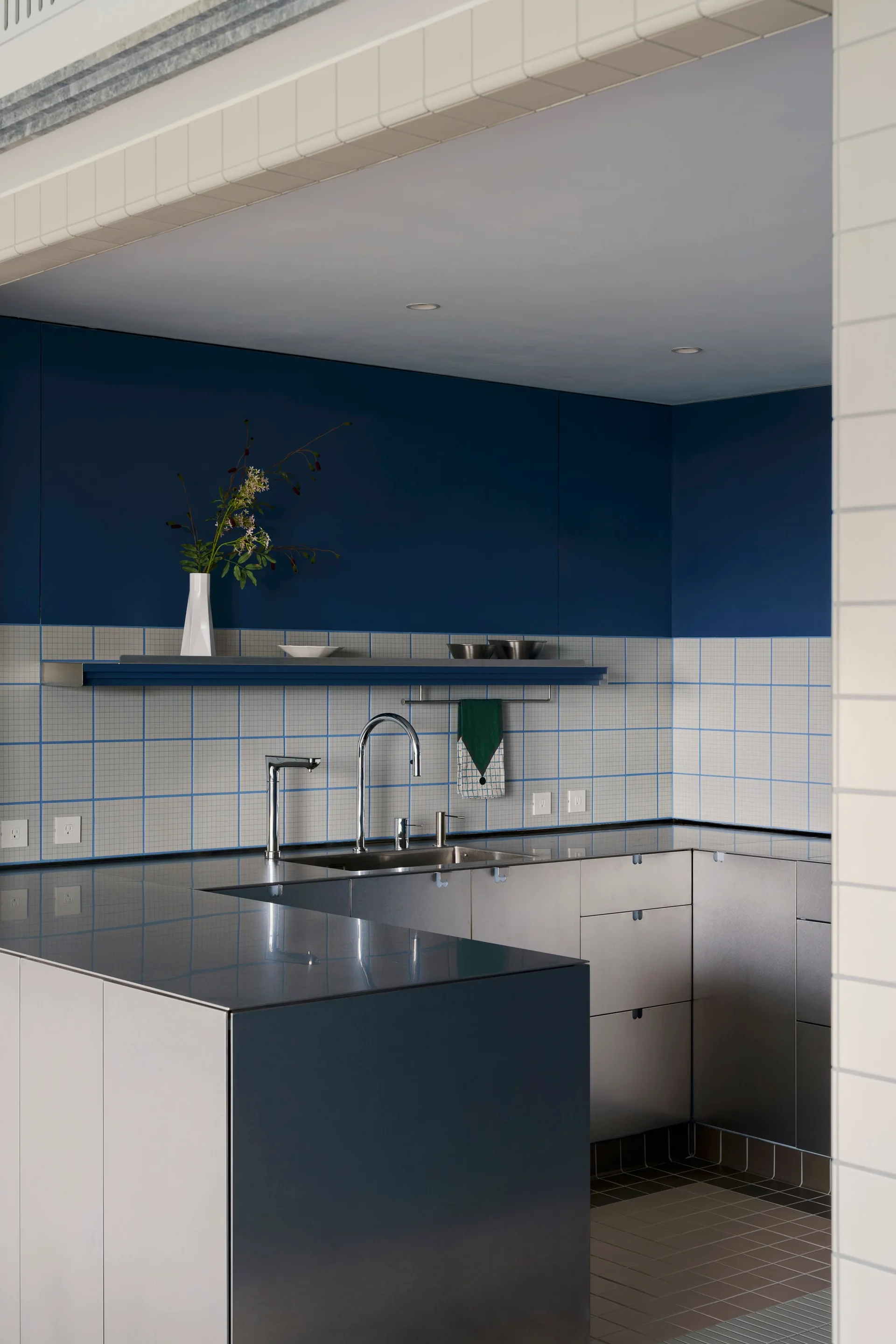 A modern kitchen features stainless steel cabinets, a grid-patterned tile backsplash, and a deep blue upper wall with a matching floating shelf.