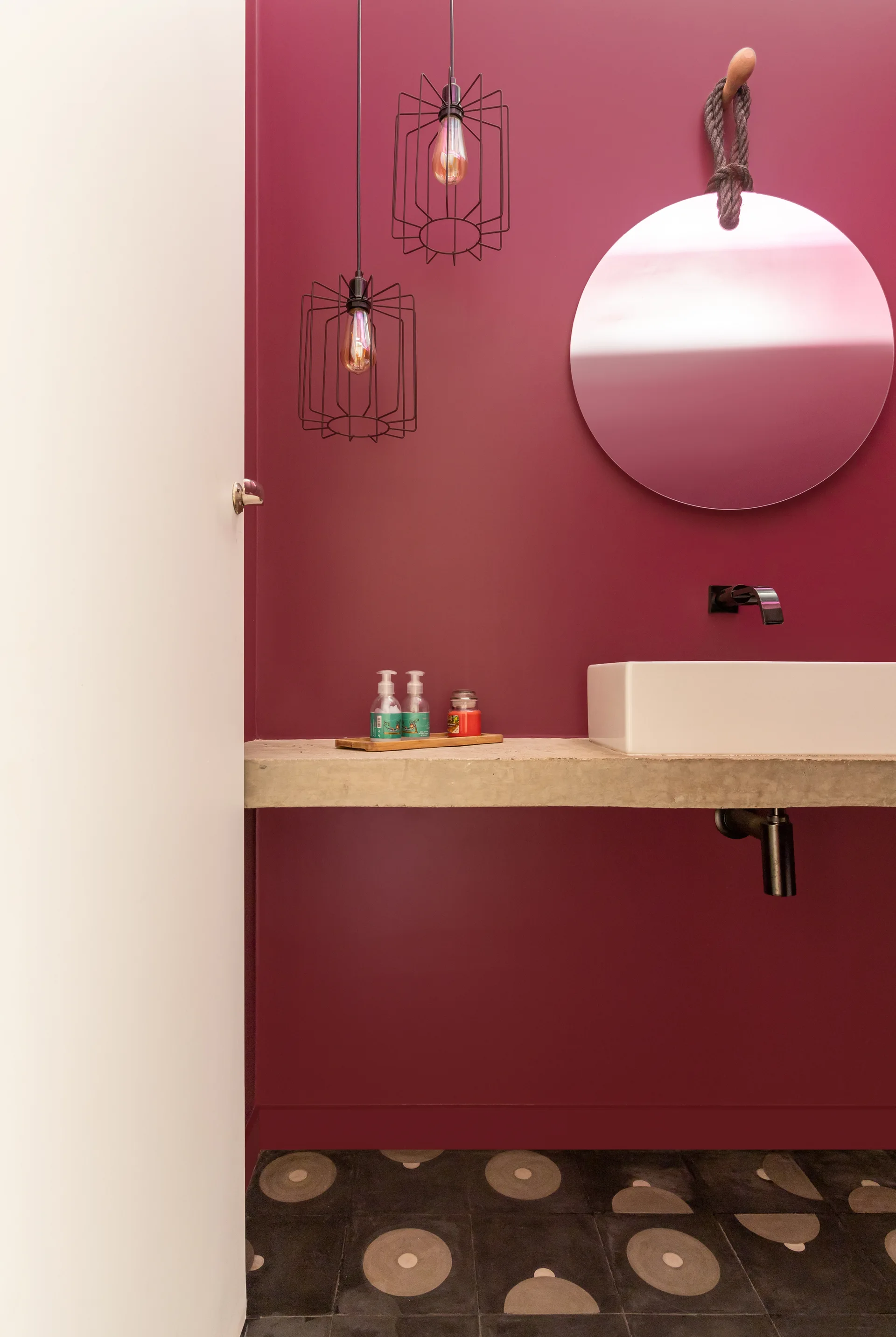 A powder room with a magenta accent wall, a floating concrete vanity with a white vessel sink, a round mirror hanging by a rope, and black cage pendant lights.