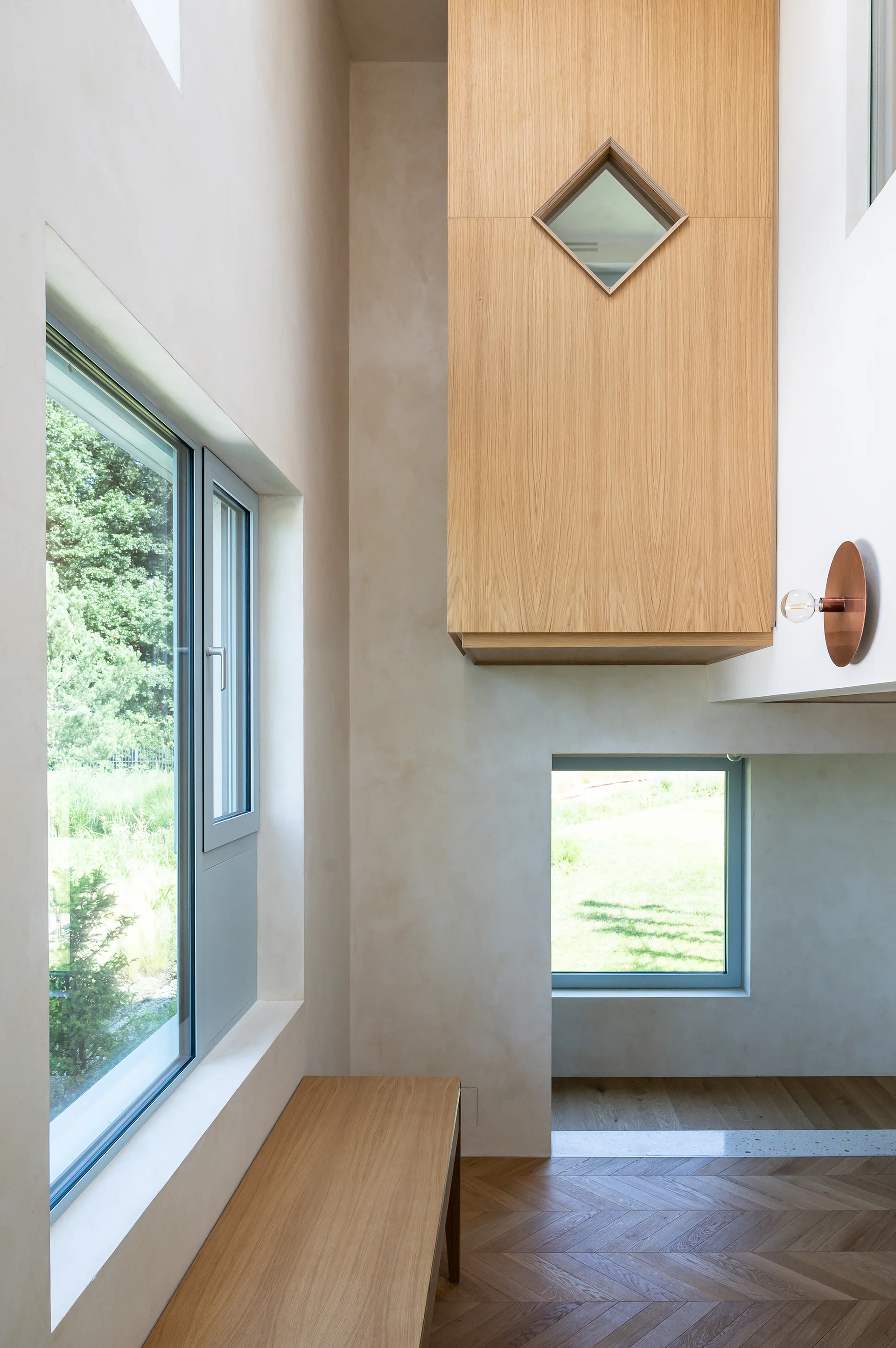 A modern hallway features herringbone wood floors, a suspended oak storage volume with a diamond window, and a matching wooden bench.