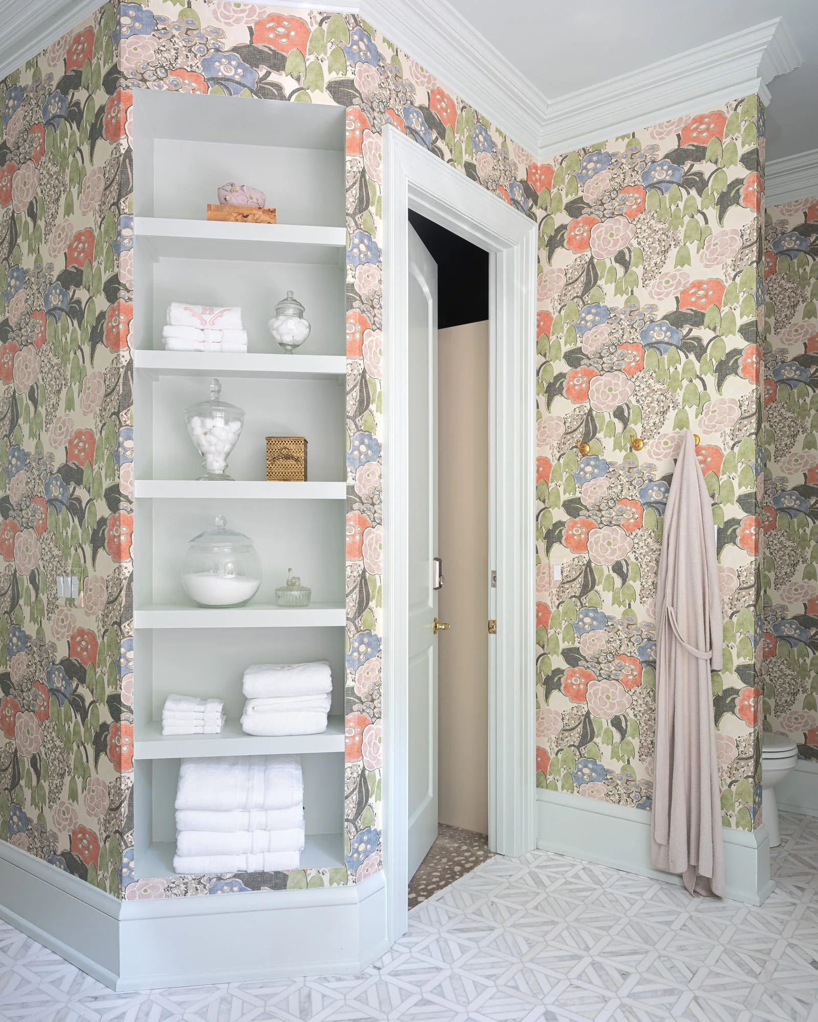 A bathroom features multi-colored floral wallpaper, white built-in shelving stocked with towels, a geometric tile floor, and a partially open door.