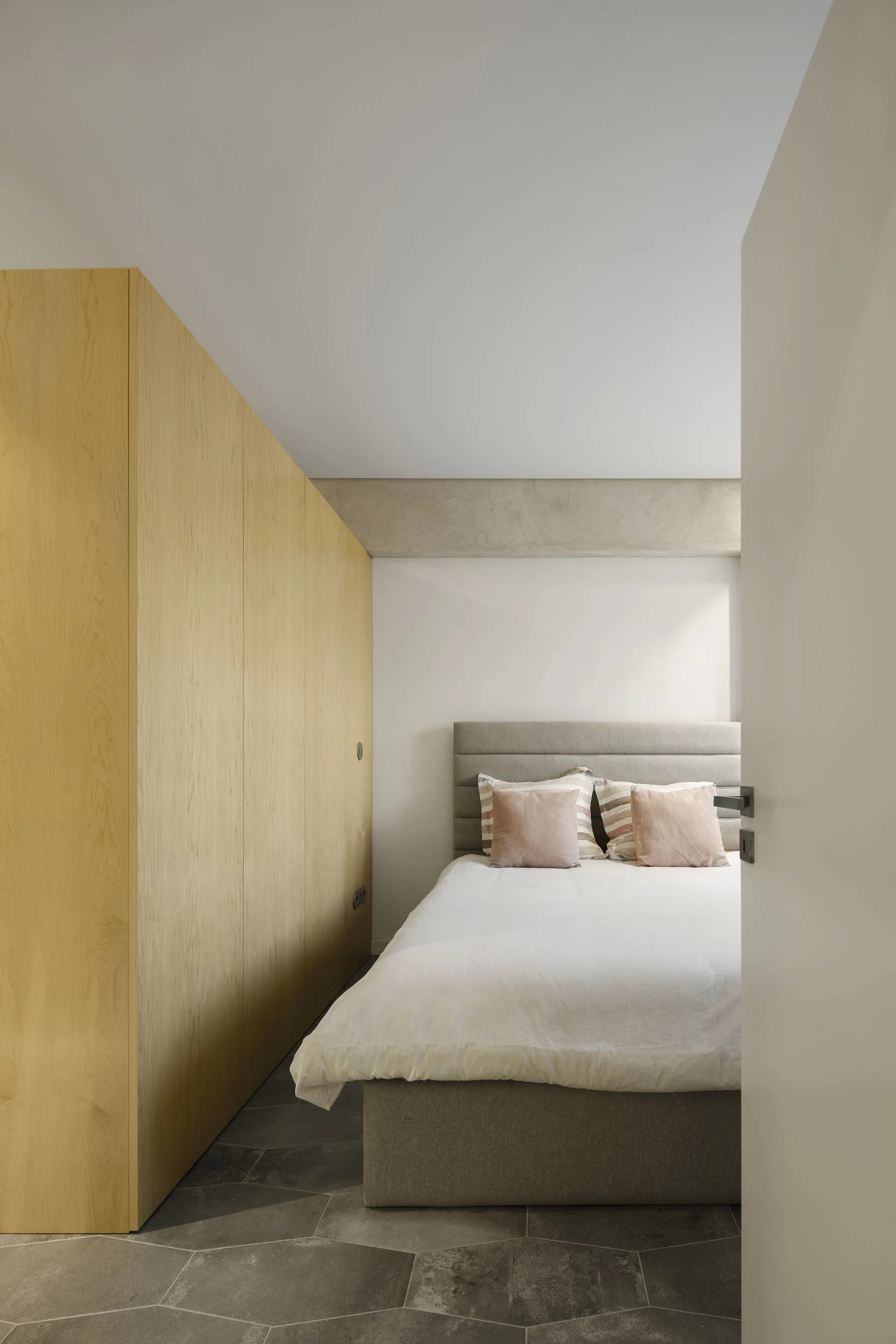 A minimalist bedroom featuring a tall light-wood wardrobe, a grey upholstered bed with pale pink pillows, and grey hexagonal tiled floors.