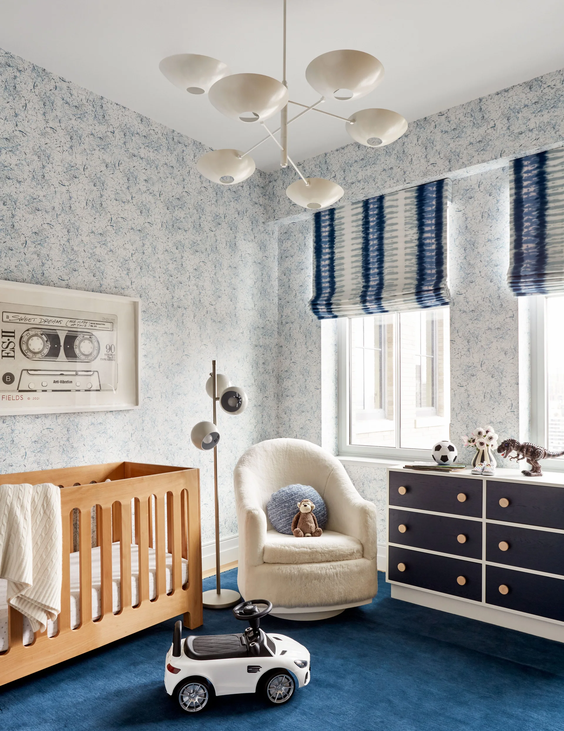 A bright nursery features a wooden crib, a white fleece armchair, a dark blue dresser, and mottled blue-and-white wallpaper.