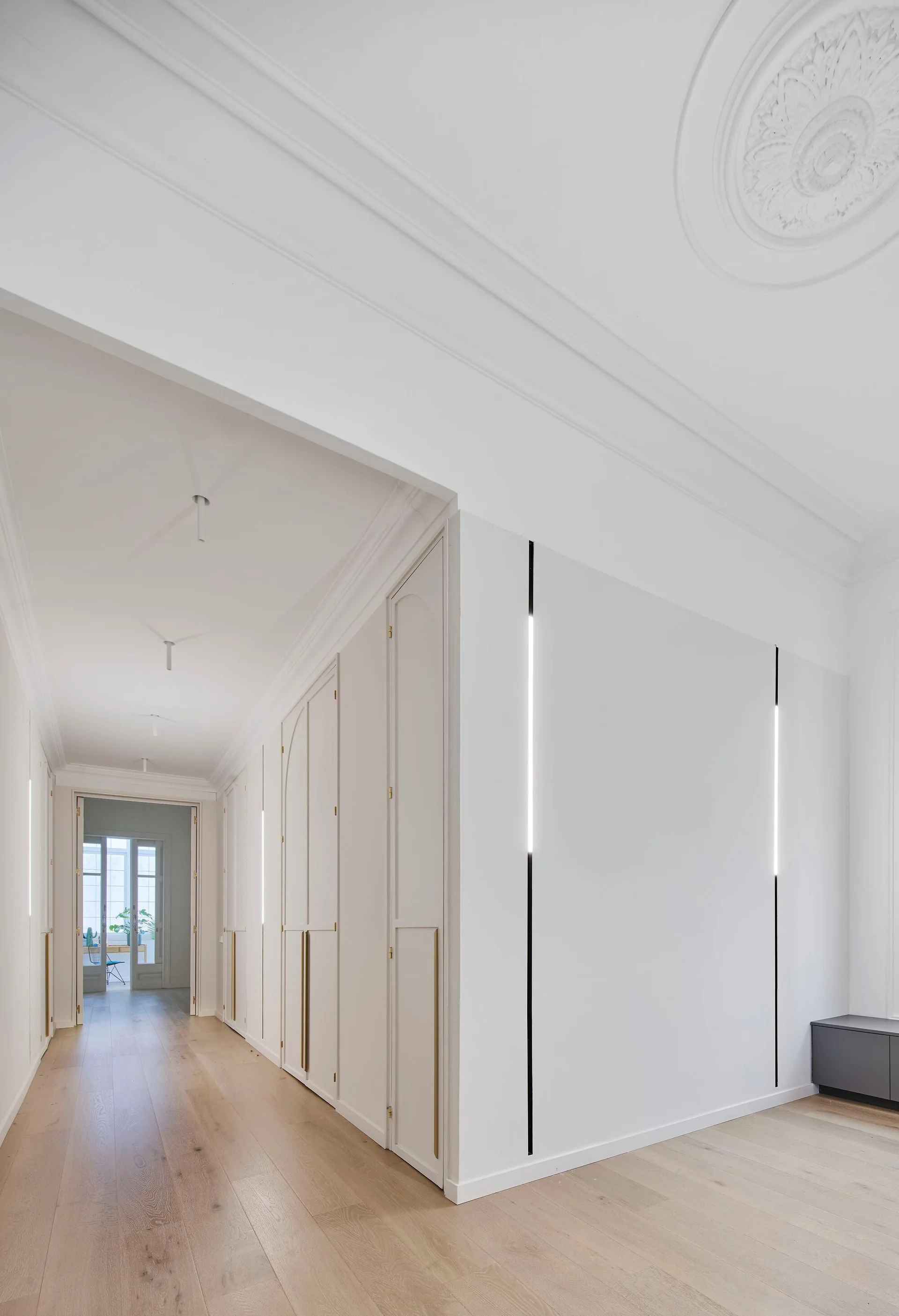 A bright white hallway with light wood floors features tall built-in wardrobes and vertical integrated LED light strips embedded in the walls.