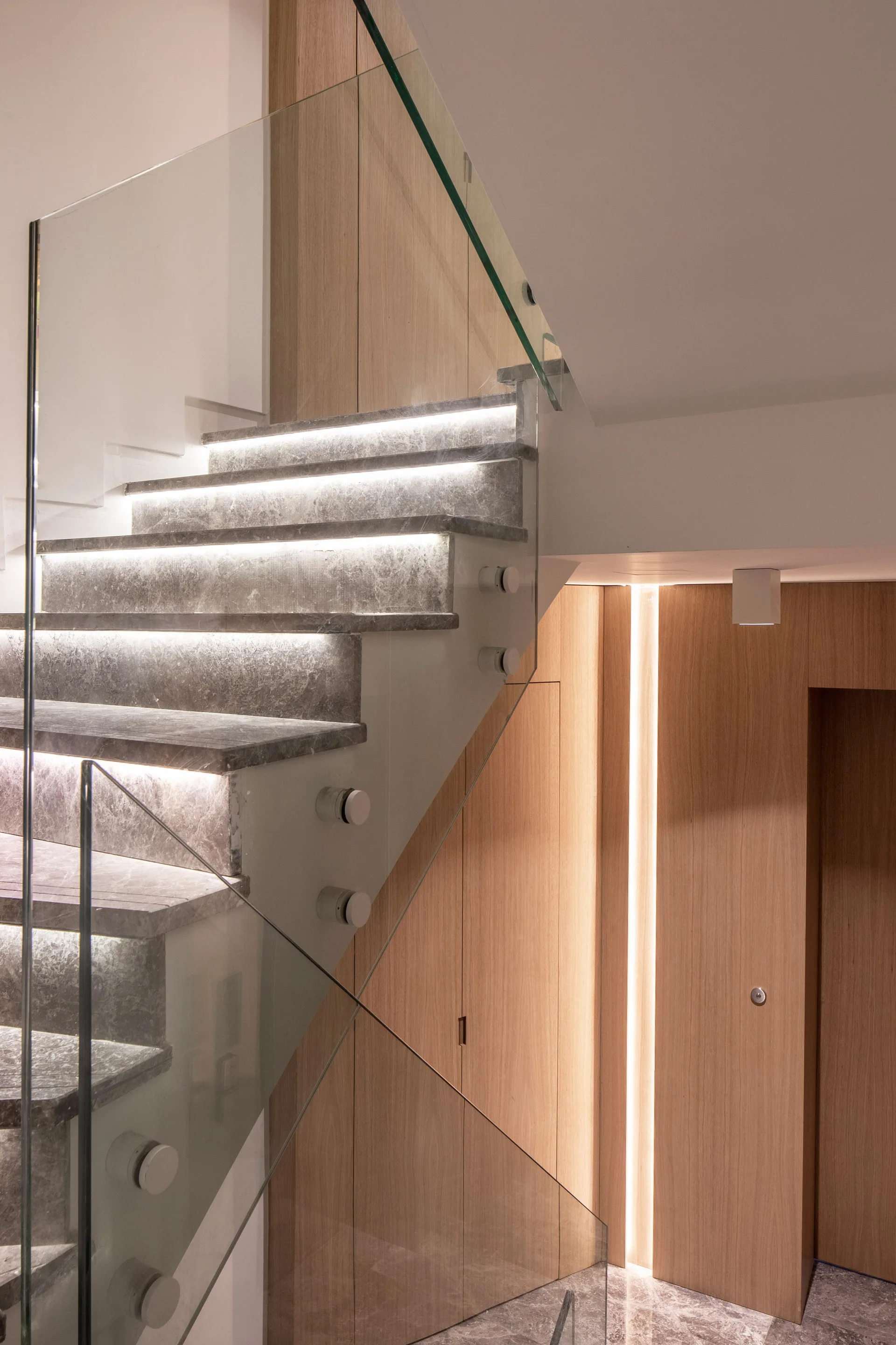 A modern staircase features grey stone steps with integrated LED lighting, a frameless glass balustrade, and light wood wall paneling.