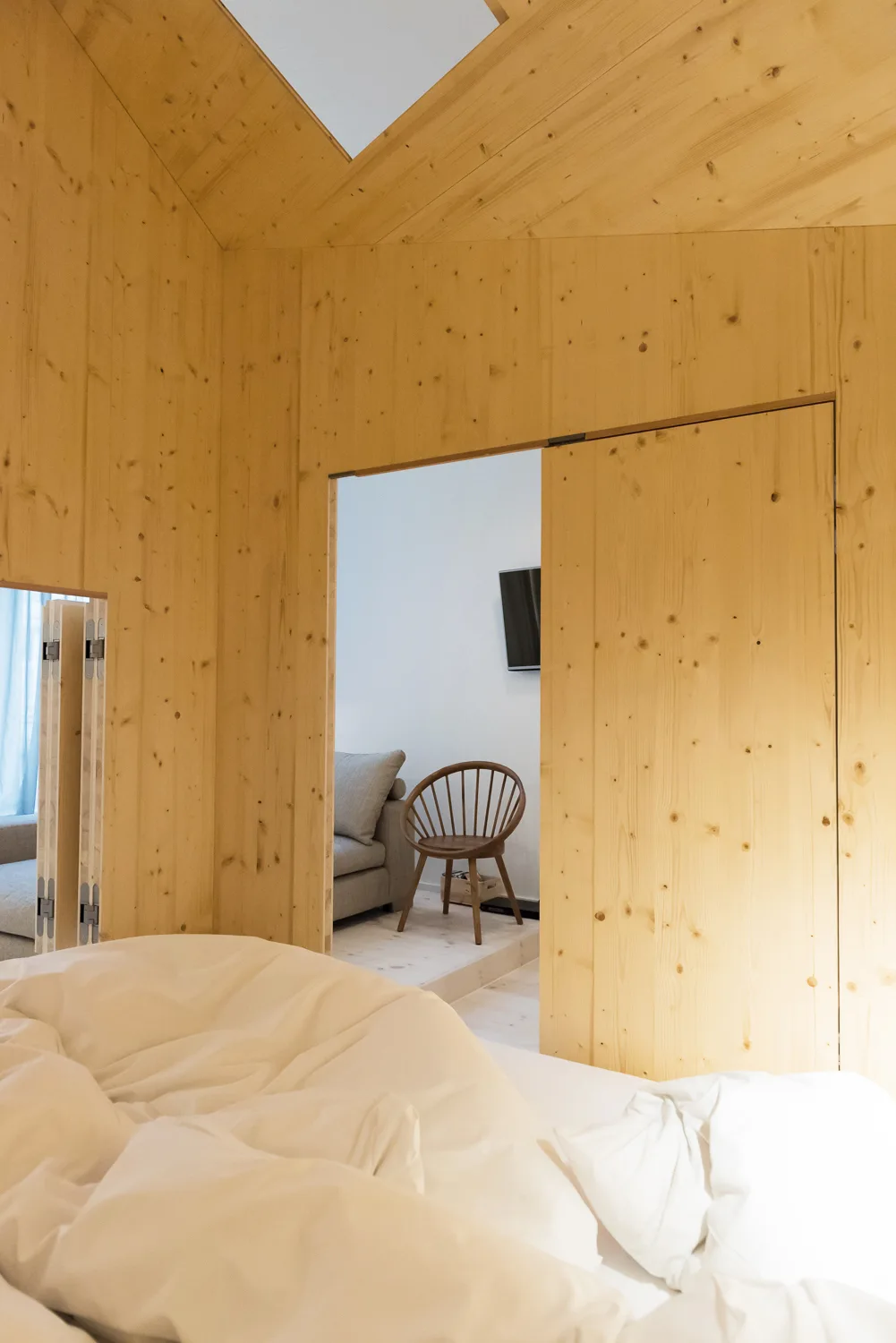 A bedroom with unmade white bedding and knotty pine wood paneled walls features a doorway opening into a sitting area with a spindle chair.