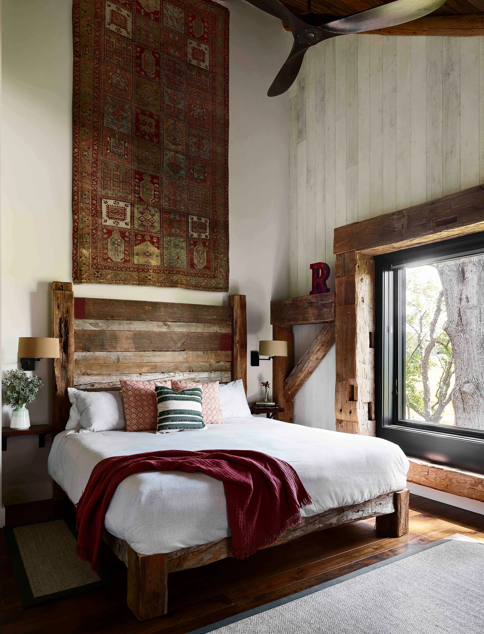 A rustic bedroom features a bed with a reclaimed wood headboard, a patterned red tapestry on the wall, and exposed timber framing.
