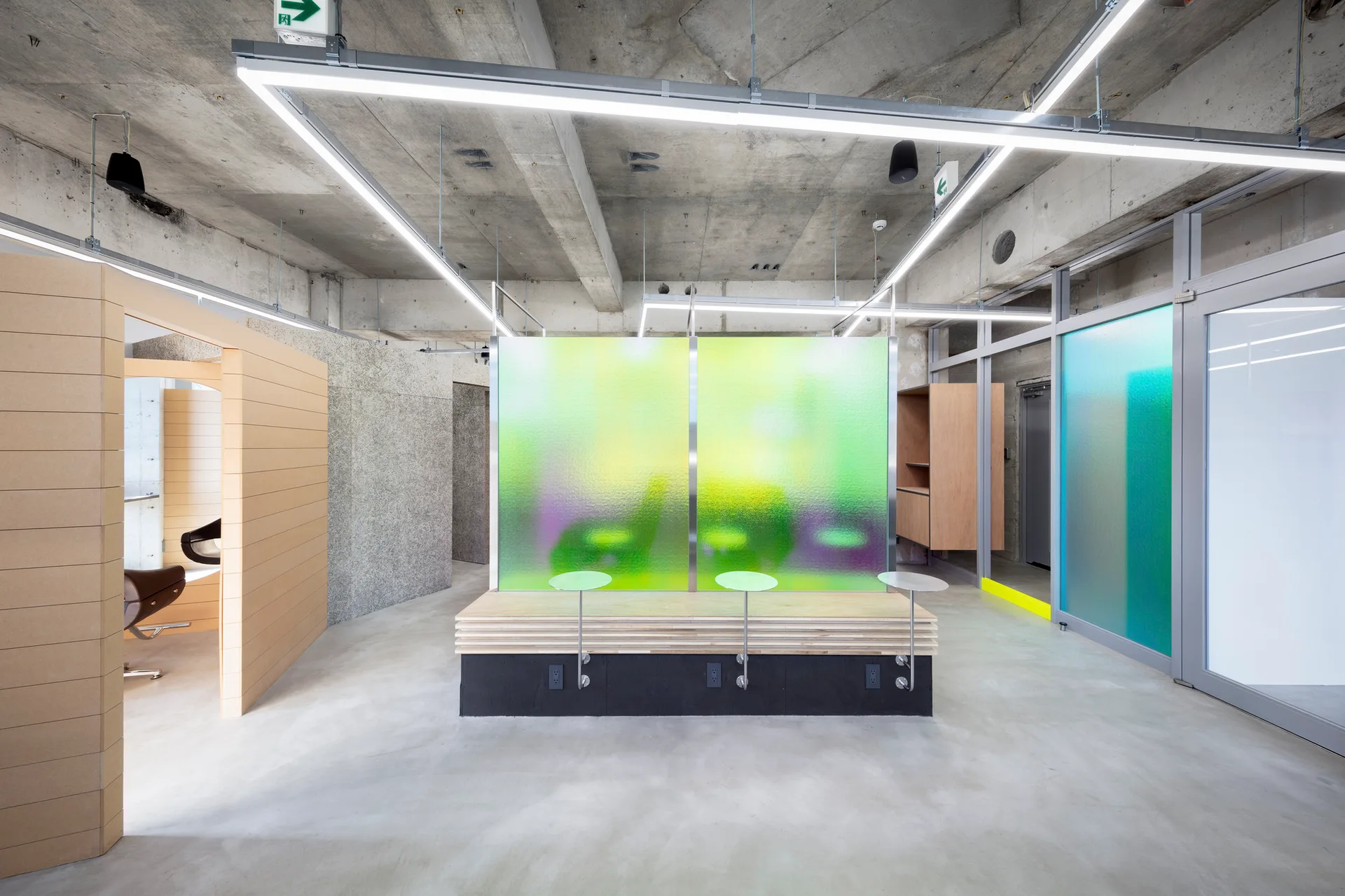 A minimalist office space with an exposed concrete ceiling, a central wooden bench, and a vibrant green and purple textured glass partition.