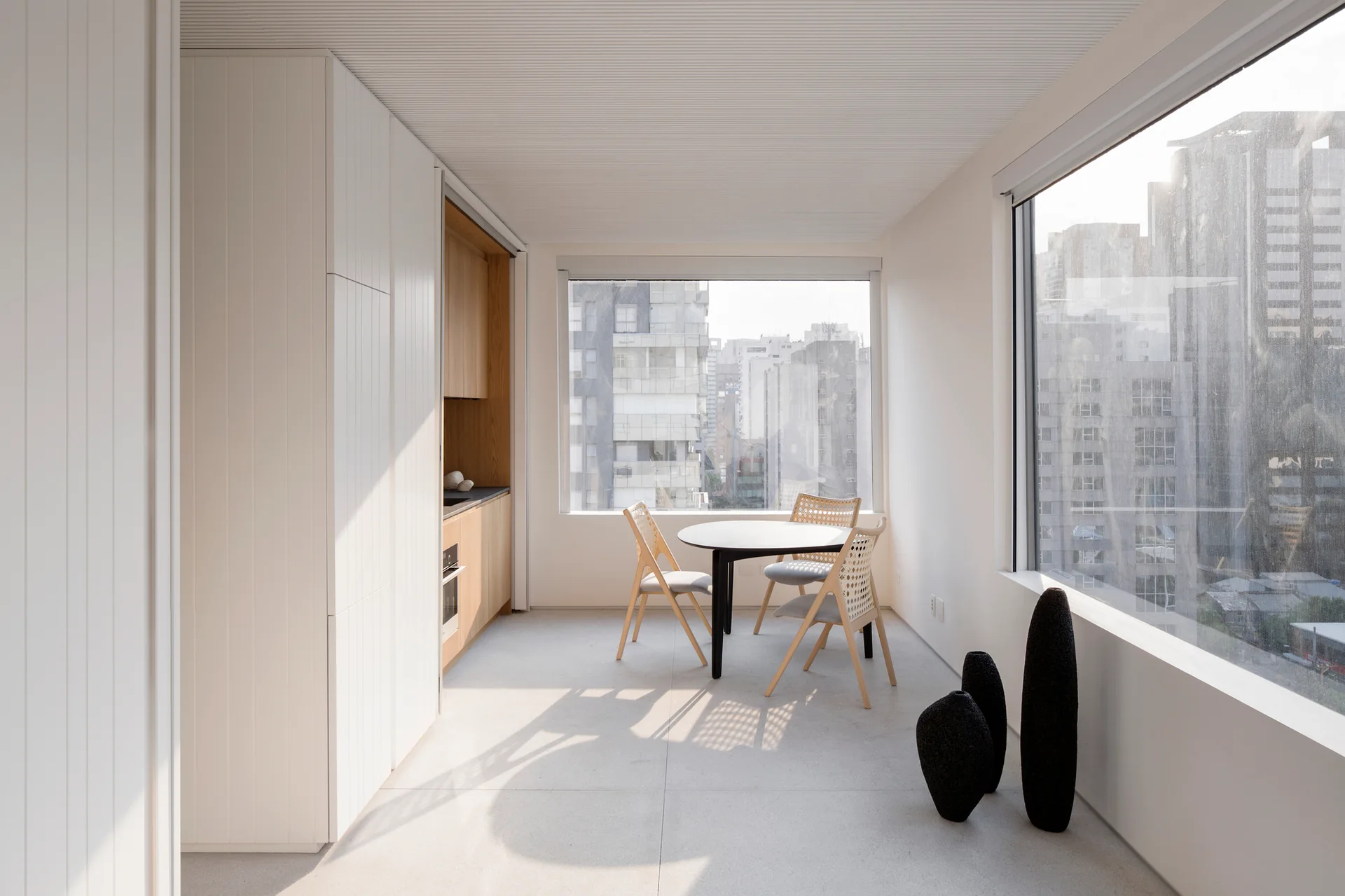 A minimalist dining room features a black round table, light wood wicker chairs, white built-in cabinets, and large windows with a city view.