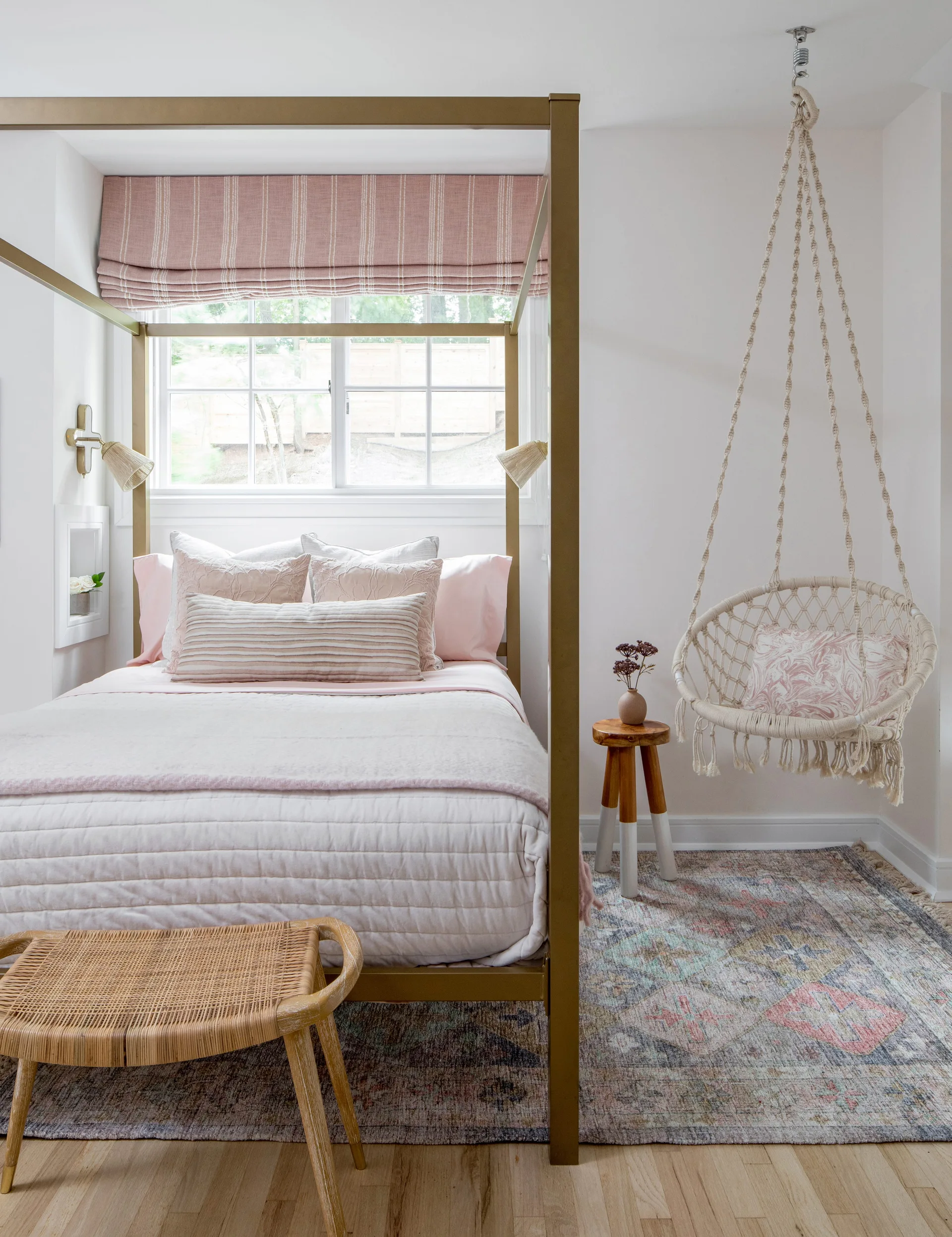 A bright bedroom features a gold canopy bed with pink bedding, a striped roman shade, and a hanging macrame chair.
