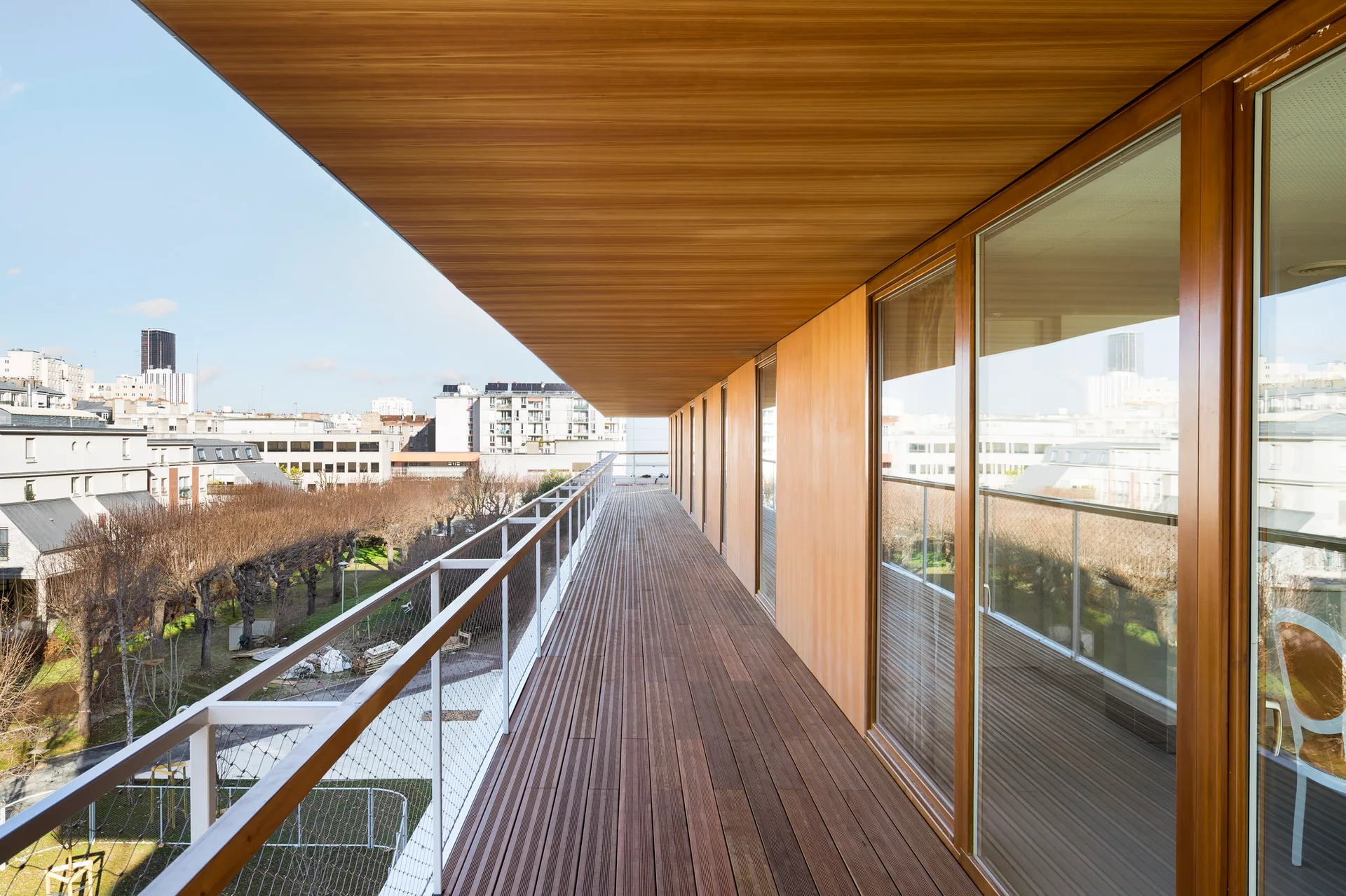 A long covered balcony features warm wood-toned decking, a matching wood ceiling, glass sliding doors, and a white metal safety railing.