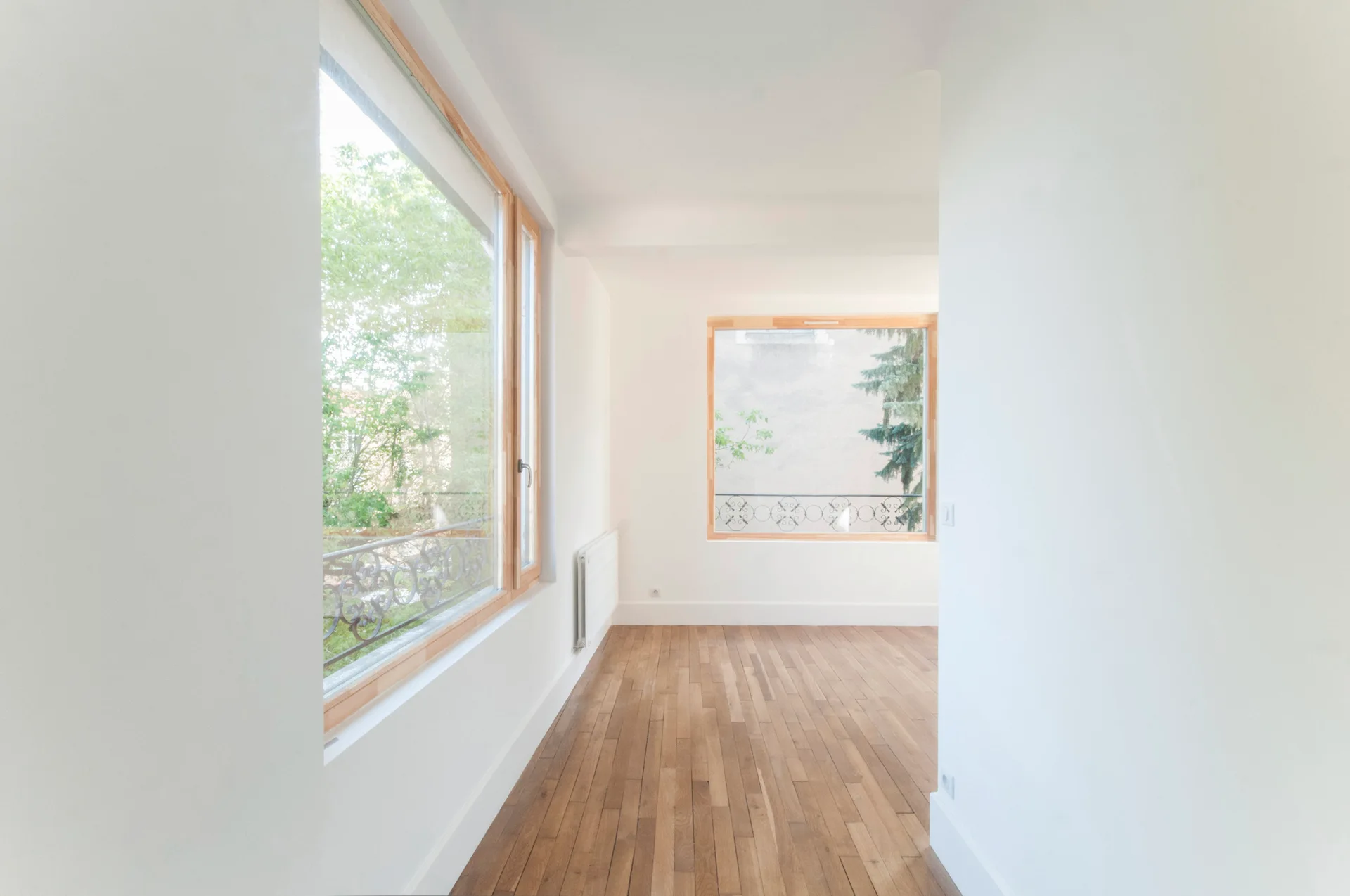 An empty room features white walls, medium-toned hardwood plank flooring, and large windows with natural wood frames looking out to green trees.