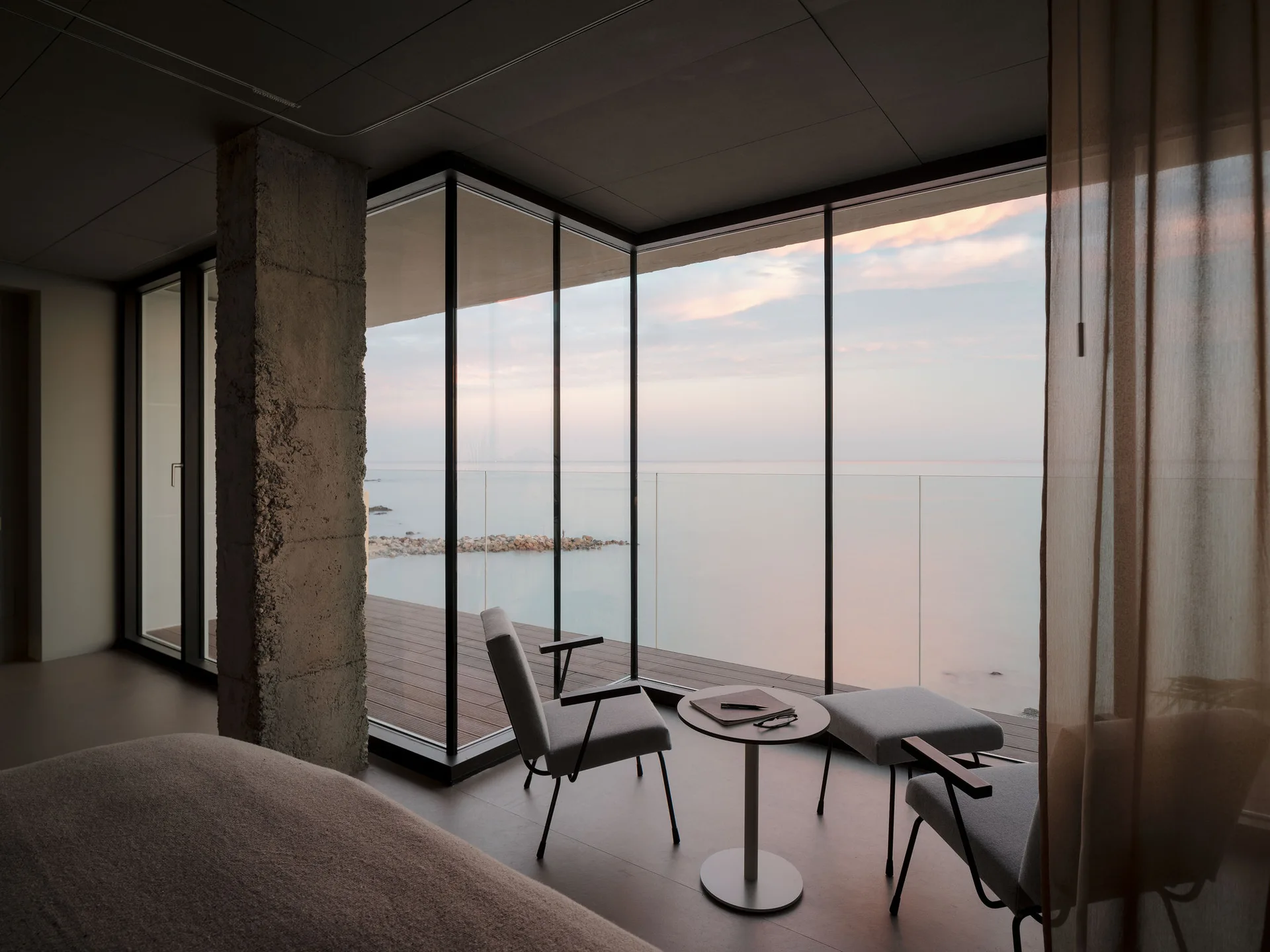 Bedroom seating area with two gray armchairs, a small table, and an exposed concrete column next to large windows overlooking the sea.
