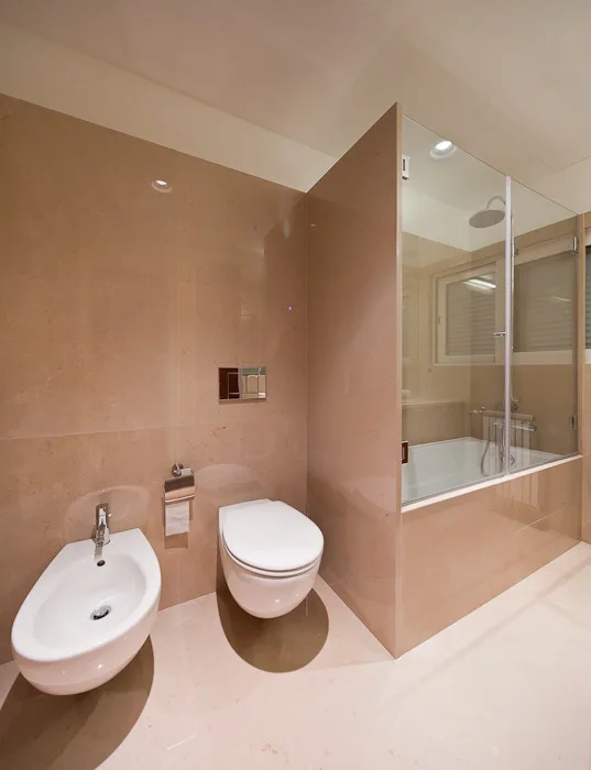 A minimalist bathroom features glossy beige stone wall cladding, a white wall-mounted toilet and bidet, and a glass-enclosed bathtub.