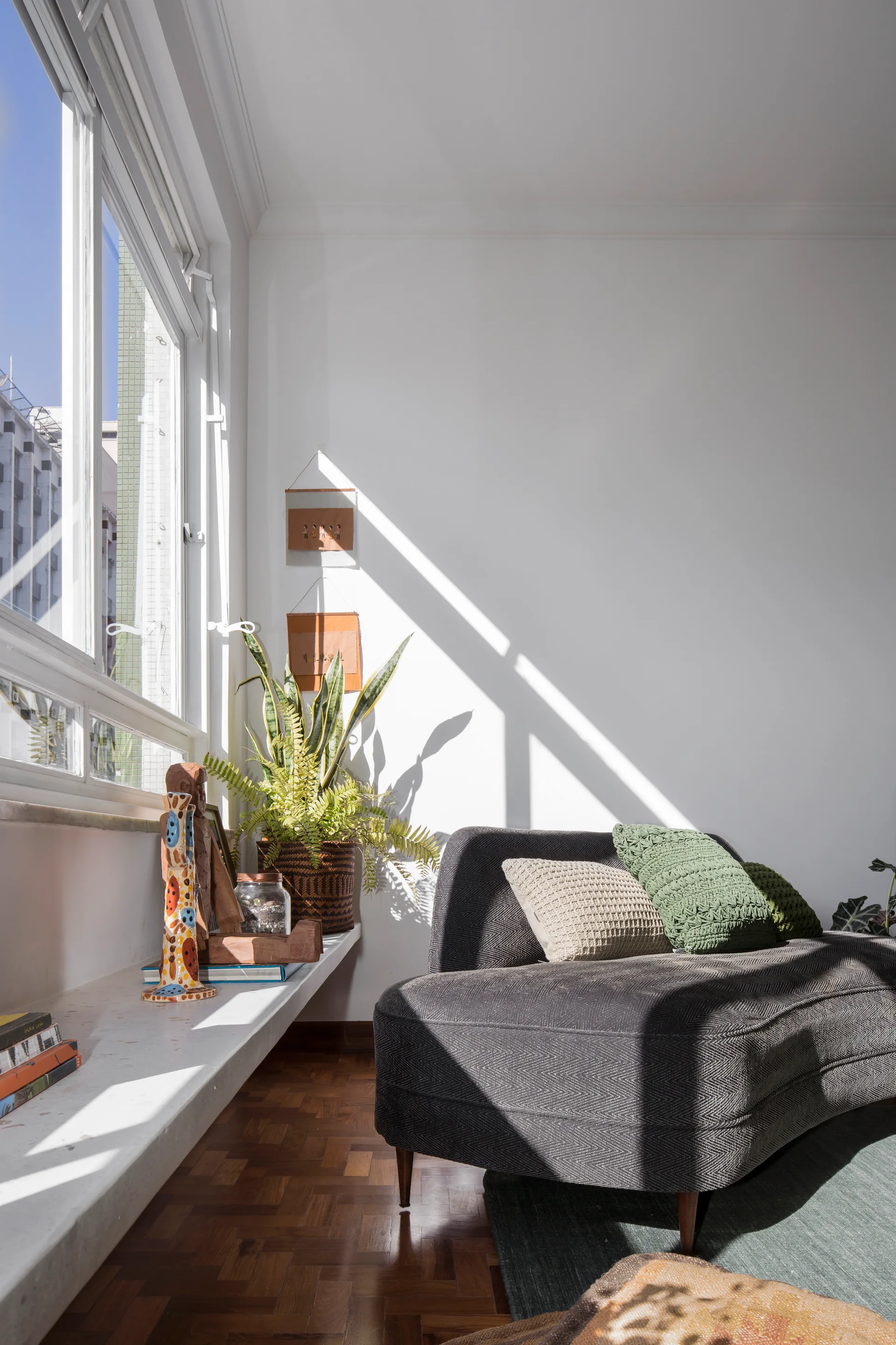 A grey chevron sofa with textured pillows sits next to a sunlit window with a long display ledge holding plants and decor.