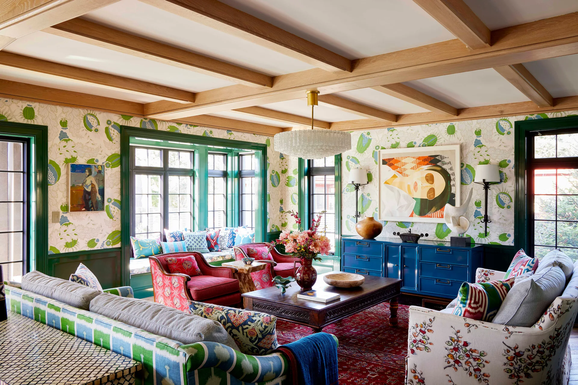 A maximalist living room featuring floral wallpaper, bright green window trim, exposed wood ceiling beams, a blue sideboard, and patterned sofas.