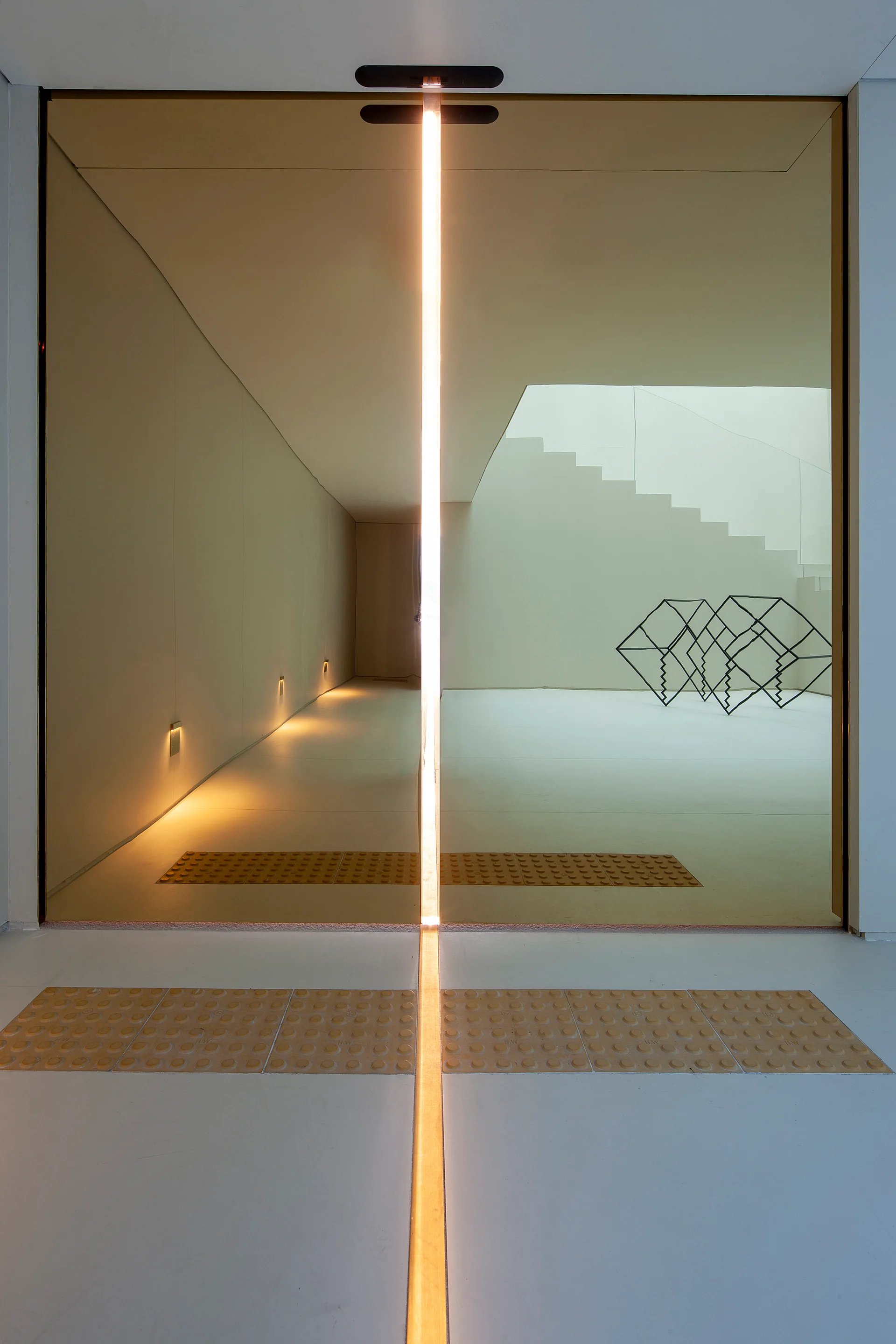 A minimalist hallway featuring a vertical glowing light strip on a glass partition, with tactile floor paving and a staircase in the background.