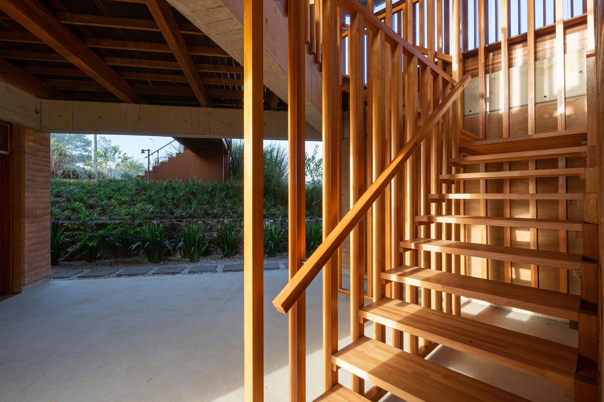 A warm wood staircase with open risers and vertical slat balustrades stands on a smooth concrete floor near an outdoor garden transition.